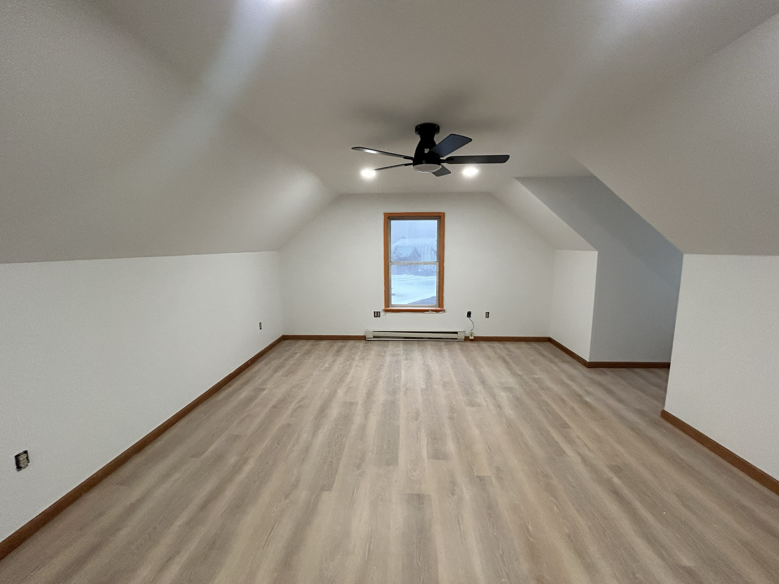 Empty attic room with sloped ceilings, a window with wooden trim, a ceiling fan, and light wood flooring.