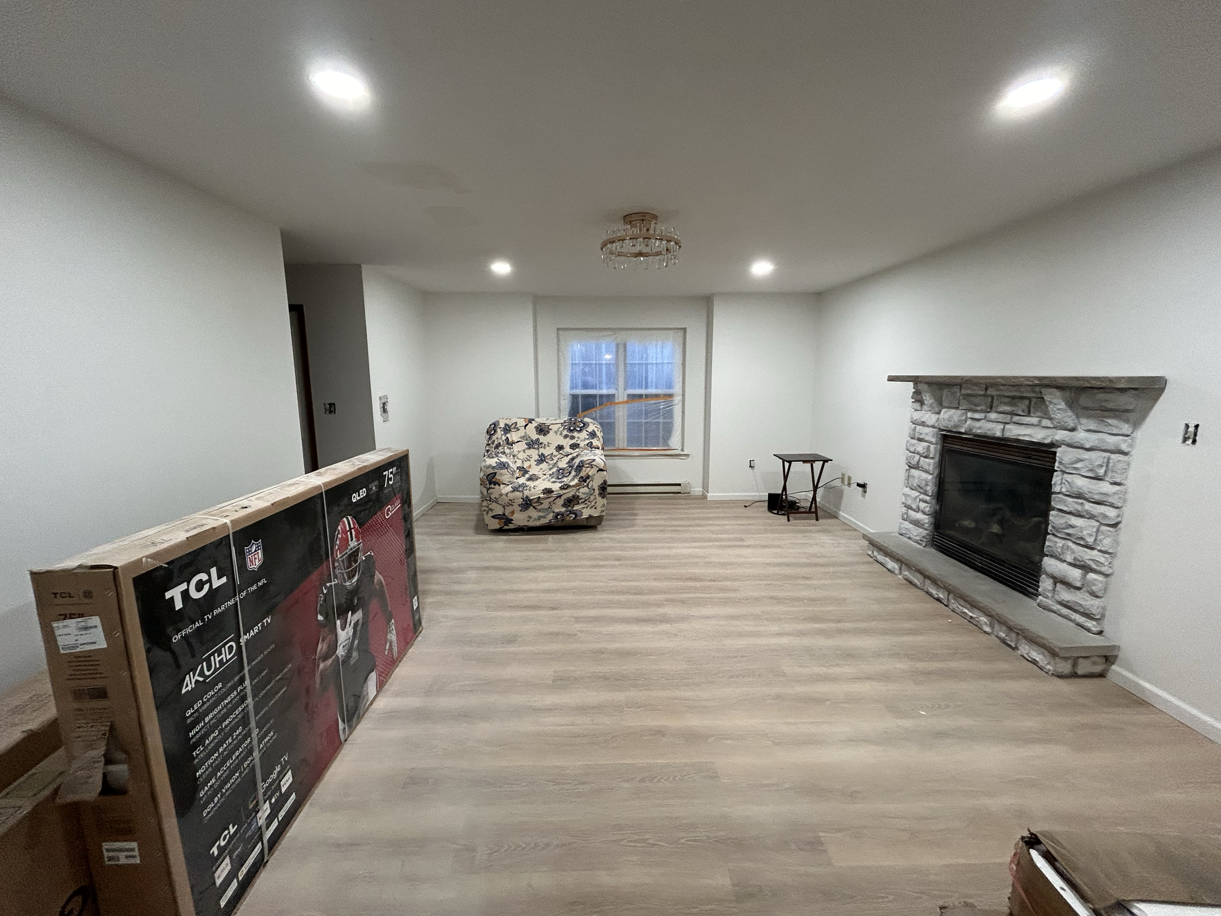 A spacious living room with a fireplace, hardwood flooring, a window, a floral armchair, a small table, ceiling lights, and a boxed TV leaning against the wall.