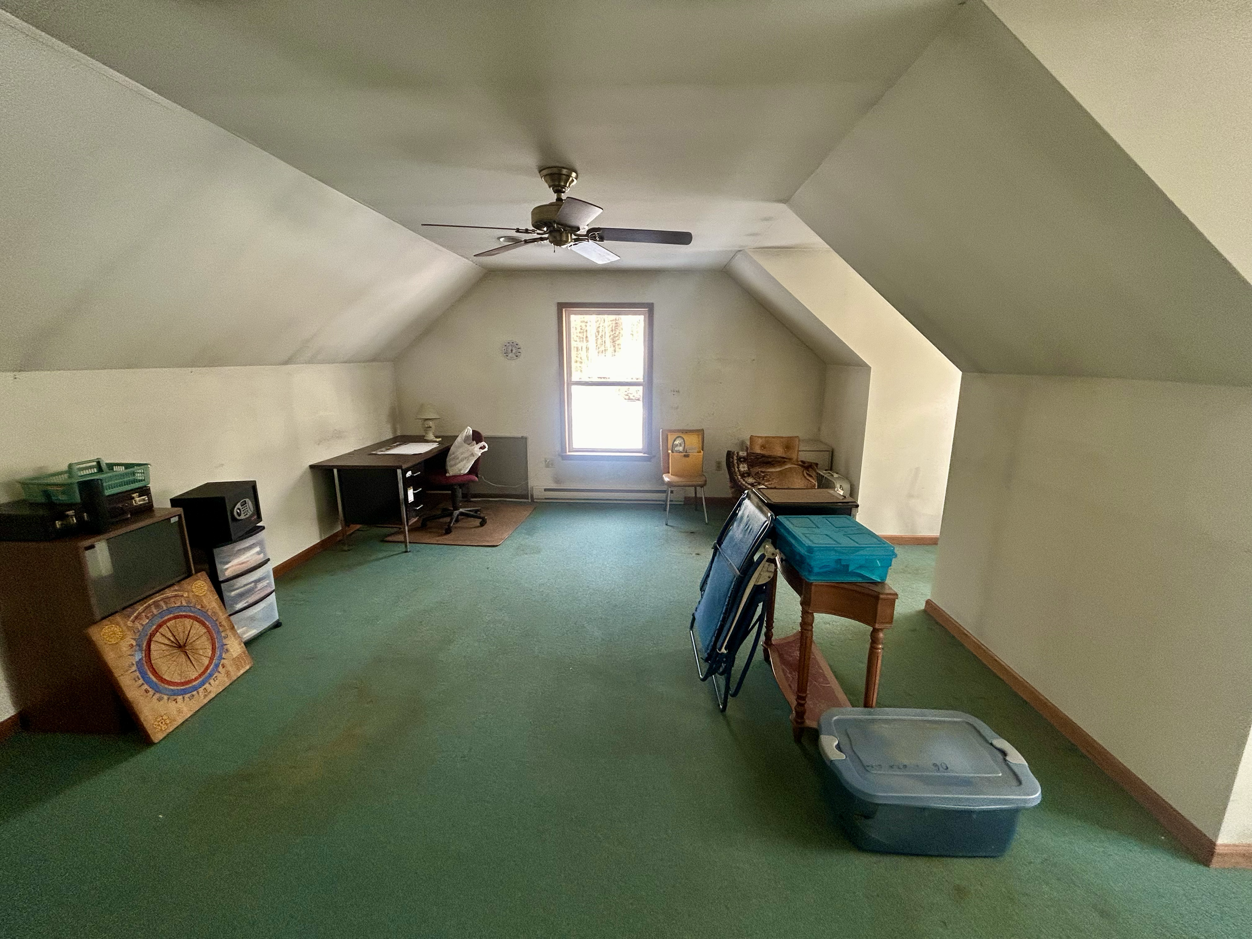 Empty attic room with green carpet, sloped ceilings, a window, ceiling fan, small desk with a chair, some stacked folding chairs, and storage containers.