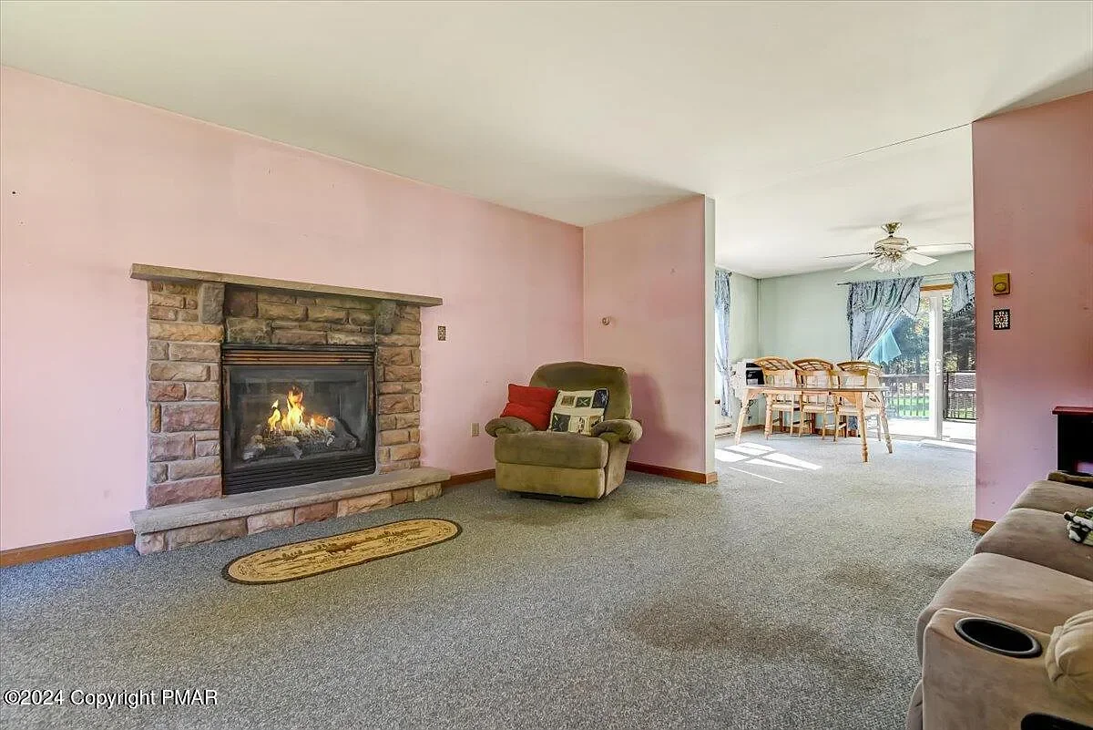 Living room with pink wall, stone fireplace, green armchair with red and patterned pillows, beige sofa with cup holder, and patterned carpeting leading to a dining area with curtains and sliding glass door.
