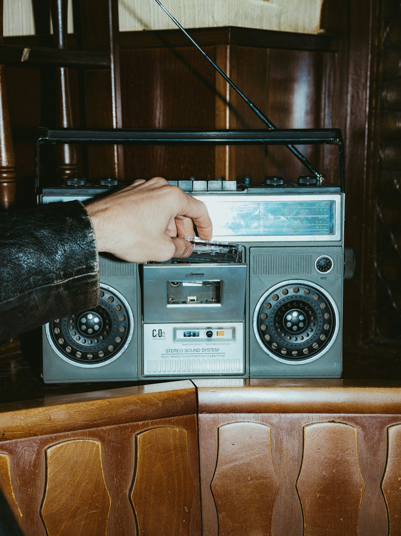 A person with a black leather jacket inserting a cassette tape into a vintage silver boombox radio on a wooden surface.