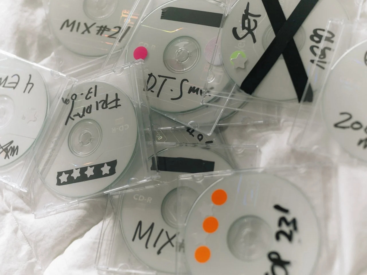 Multiple CDs in transparent plastic cases with handwritten labels and stickers, some with black tape, orange dots, and star designs, scattered on a white surface.