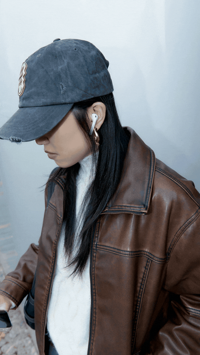 A person wearing a casual denim baseball cap, white t-shirt, and brown leather jacket. The person has long black hair, earrings, and their face is partially obscured by the cap. They are looking down and holding a smartphone.