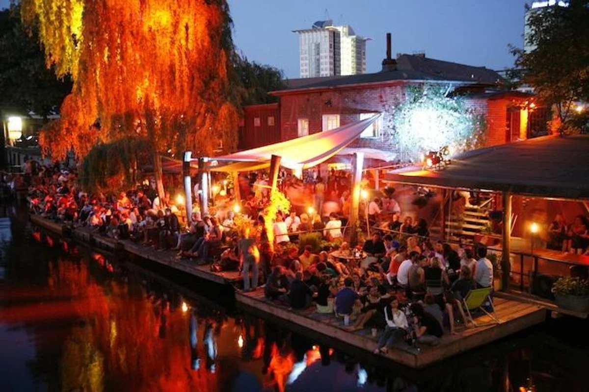 Abend in einer belebten Bar am Wasser mit vielen Gästen, Lichter, Bäumen und einem Stadt-Hintergrund.