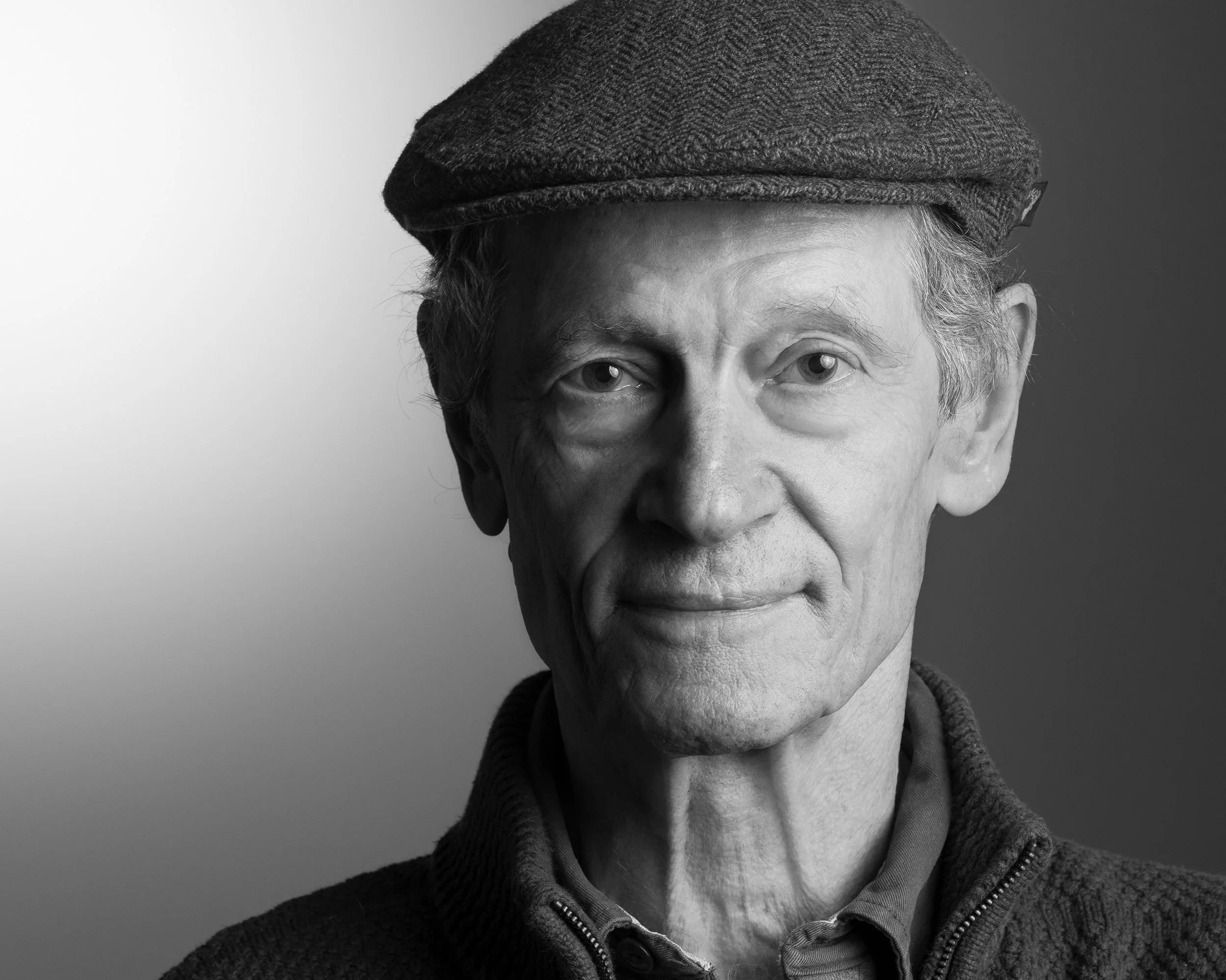 Schwarz-Weiß-Porträt eines älteren Mannes mit Mütze, mit ernstem Gesichtsausdruck, vor einem zweigeteilten dunklen und hellen Hintergrund.