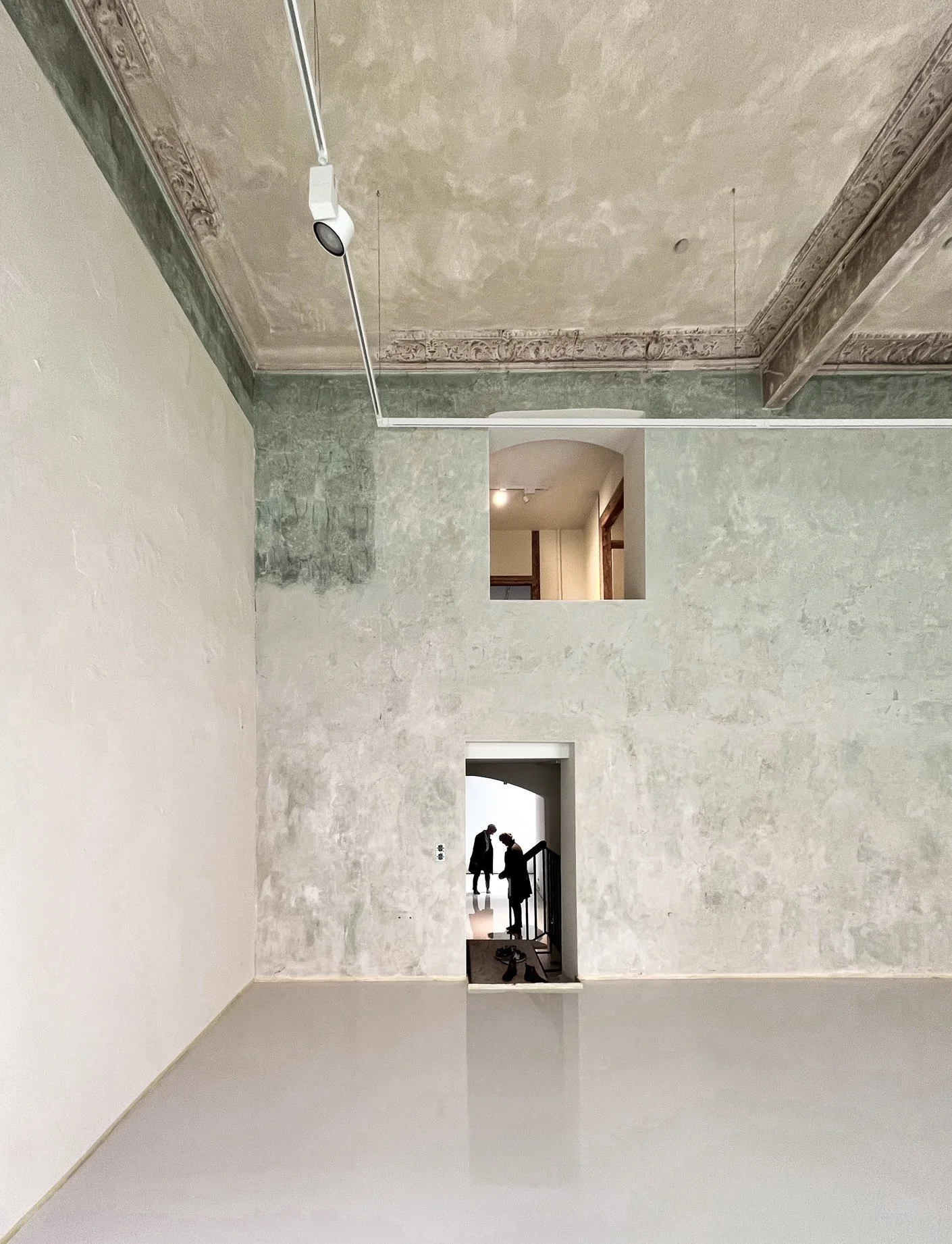 Ein leeres, historisches Raum mit hohen Wänden und Decke, während im Hintergrund Menschen in einem Museum oder einer Galerie zu sehen sind