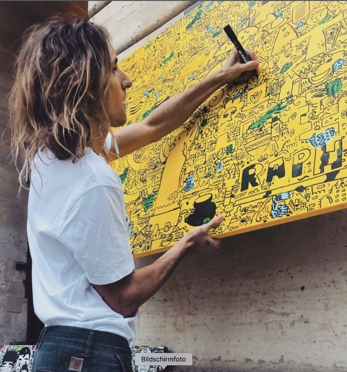 Ein Mann mit langen, gewellten braunen Haaren zeichnet auf eine große gelbe Wand mit bunten Linienstiften. Die Wand zeigt ein komplexes, detailliertes Kunstwerk mit vielen kleinen Figuren und Elementen. Der Mann trägt ein weißes Hemd und dunkle Hosen.