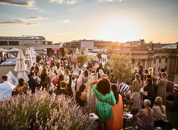 Menschen versammeln sich auf einer Dachterrasse während des Sonnenuntergangs, einige sitzen, andere stehen, mit Gebäuden im Hintergrund.