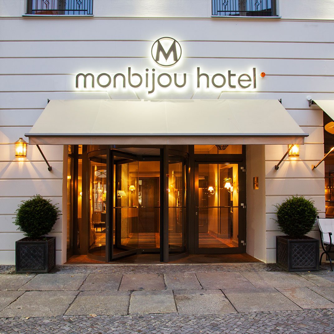 Das Bild zeigt den Eingang des Monbijou Hotels mit beleuchtetem Schild, einer runden Eingangstür, seitlichen Lampen und zwei kleinen Büschen in quadratischen Töpfen auf beiden Seiten der Tür.