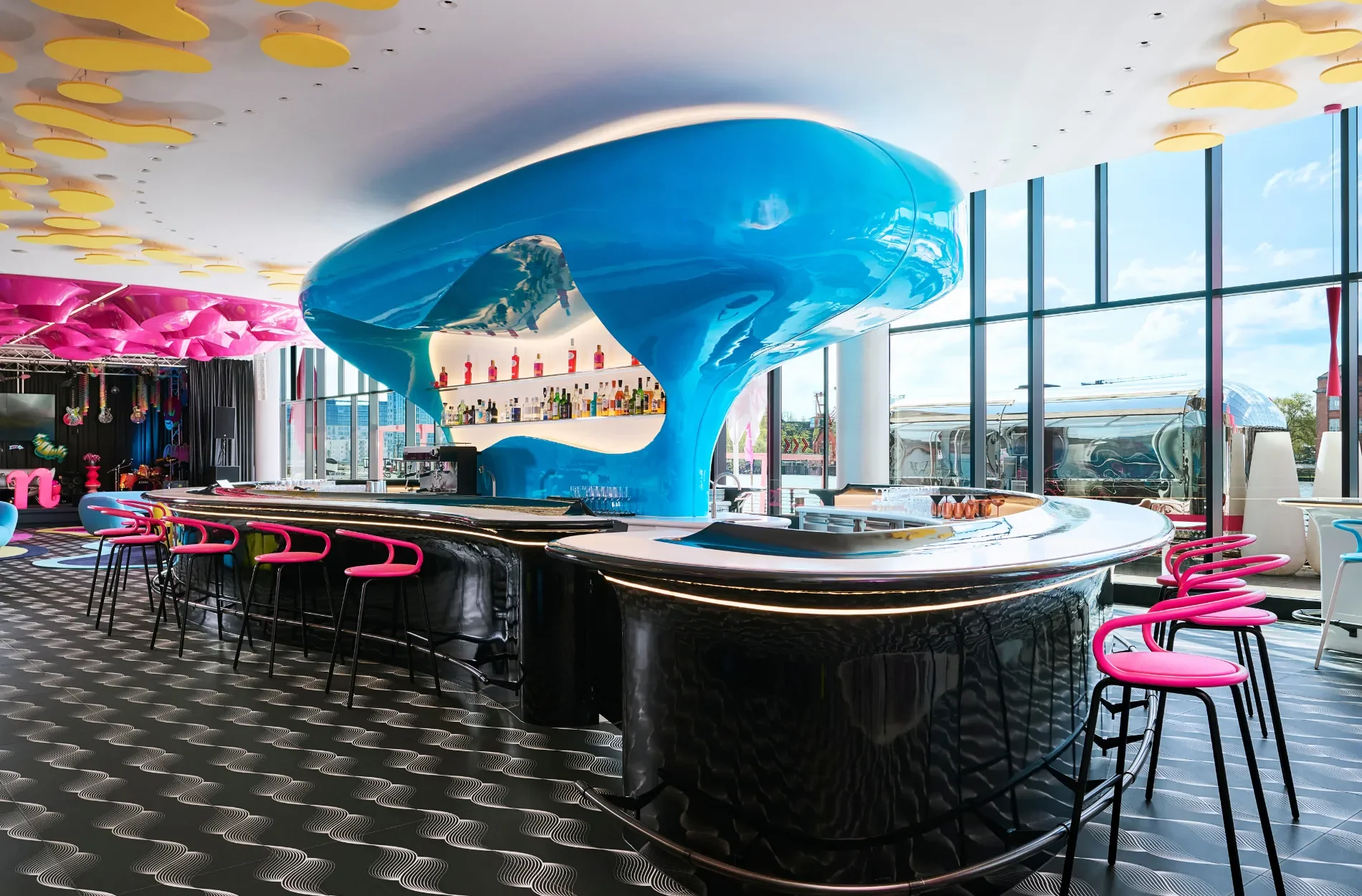 Moderner Bar mit schwarzer Theke, pinken Barhockern und einer blauen, geschwungenen Bar-Architektur, große Fenster mit Stadtblick, bunte Dekorationen und stilvolle Beleuchtung.