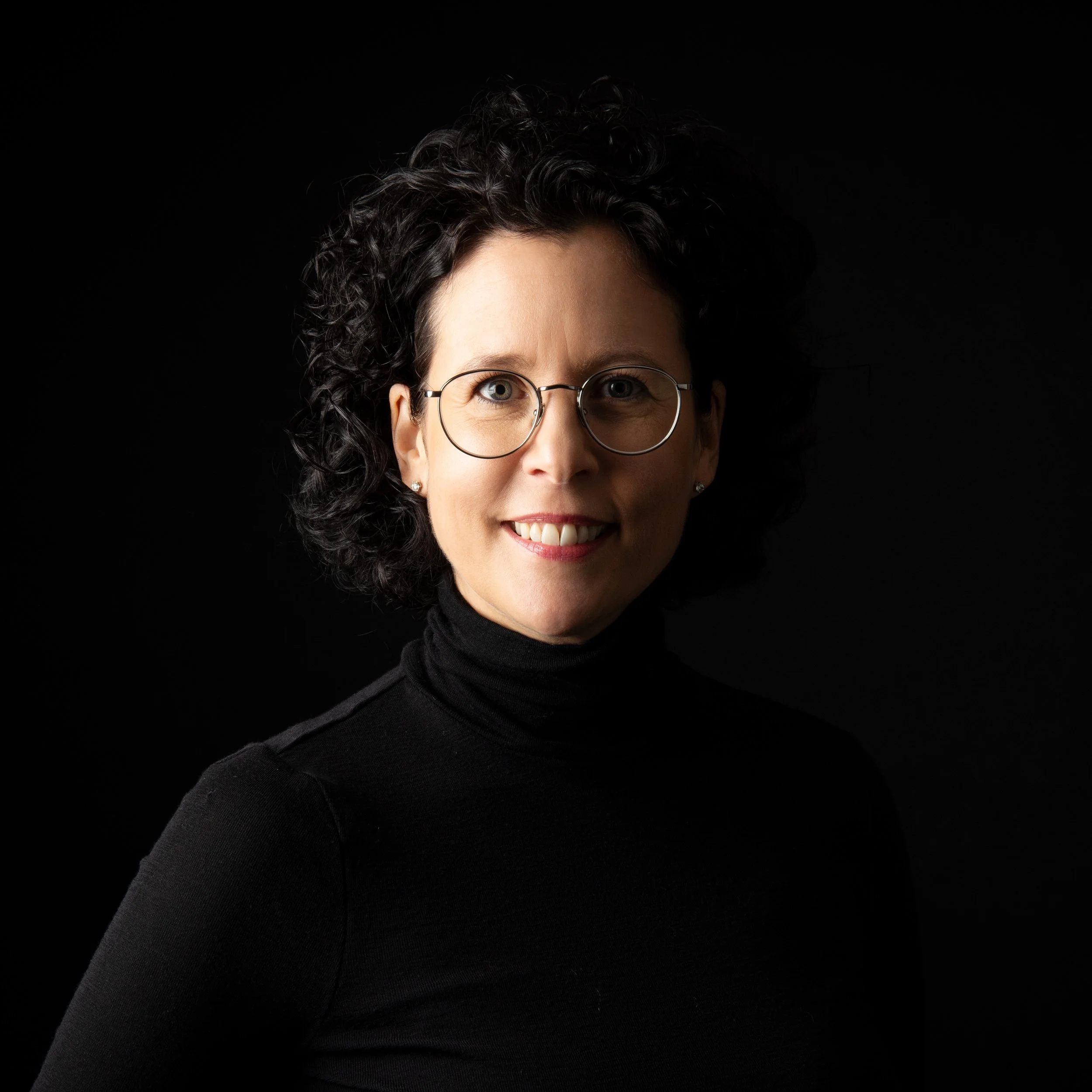 Porträt einer lachenden Frau mit dunklen, lockigen Haaren, runden Brillen und schwarzem Turtleneck vor schwarzem Hintergrund.