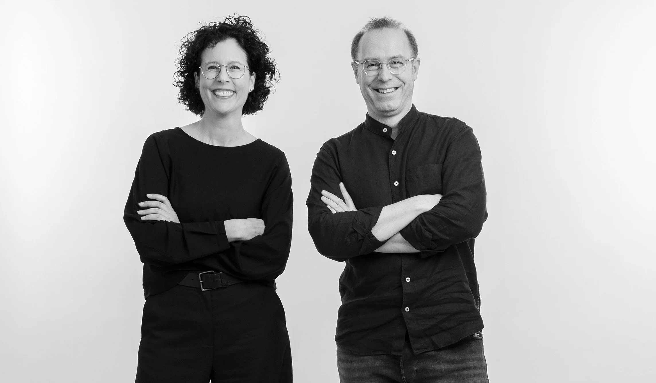 Ein Mann und eine Frau stehen nebeneinander vor einem weißen Hintergrund, lächelnd mit verschränkten Armen, in Schwarz-Weiß-Fotografie.