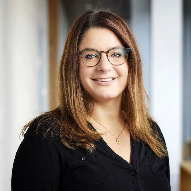 Ein junges Mädchen mit braunen, mittellangen Haaren und Brille, die lächelnd in die Kamera schaut, im Hintergrund eine helle Innenraum.