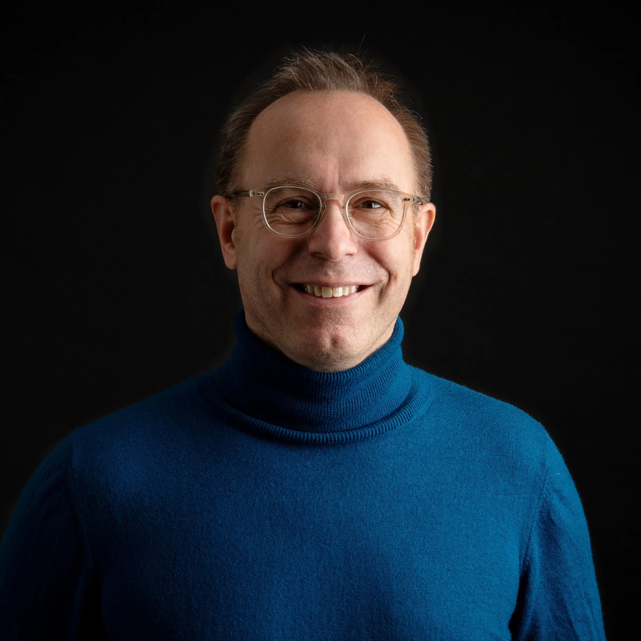Porträt eines lachenden Mannes mit Brille, blauer Rollkragenpullover, vor schwarzem Hintergrund.