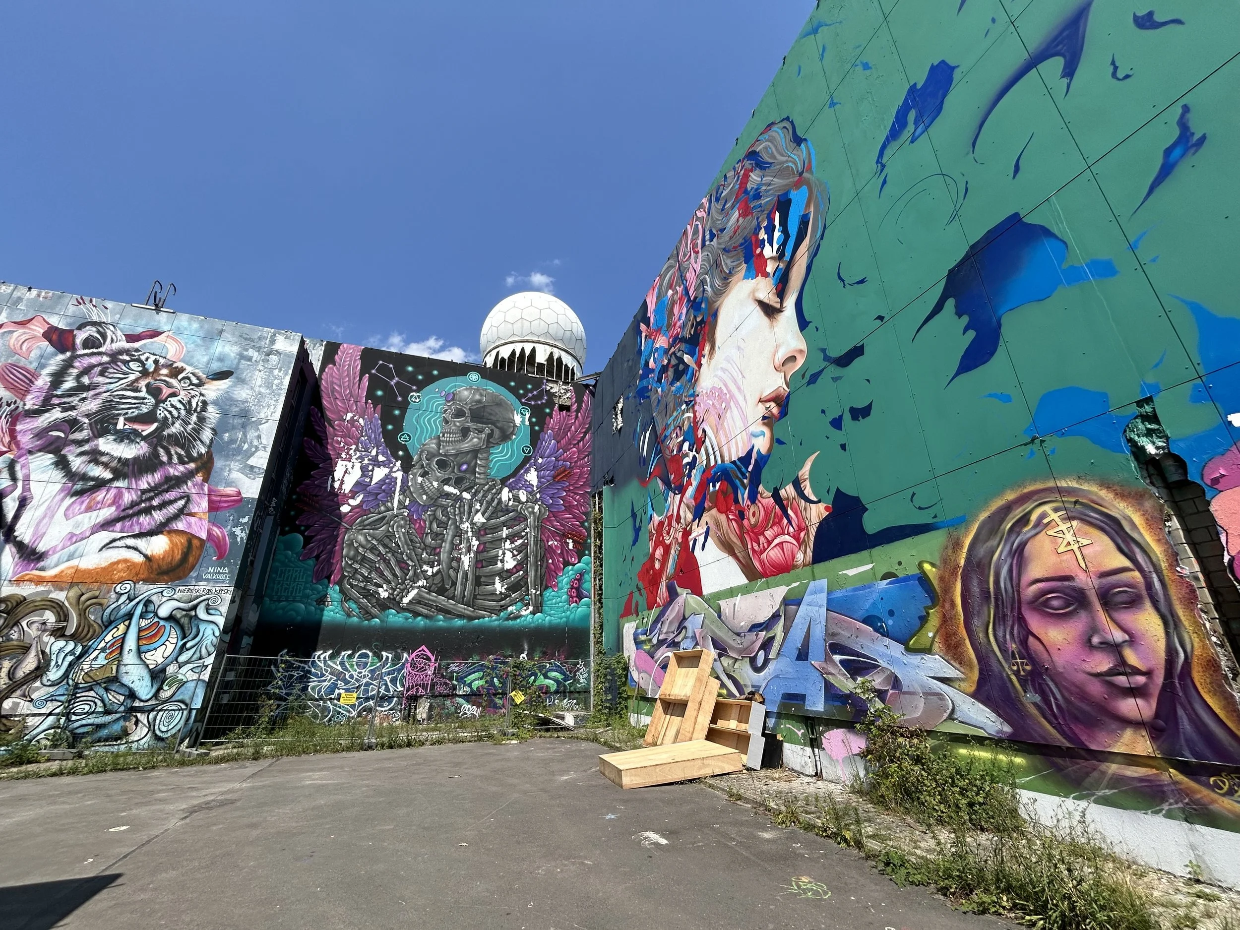 Farbenfrohe Wandmalereien mit Tieren, einer skelettähnlichen Figur, einer Frau mit blauen Haaren, und einer Frau mit mystischer Symbolik im Gesicht. Himmel im Hintergrund.