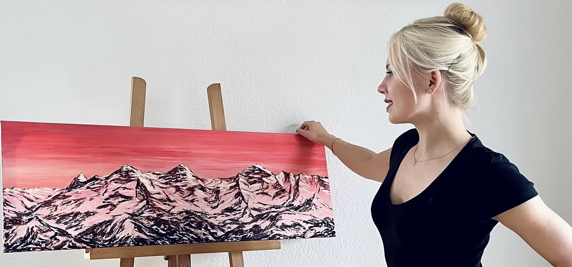 Eine Frau mit blonden Haaren in einem Dutt betrachtet ein Gemälde von schneebedeckten Bergen vor einem rosafarbenen Himmel.