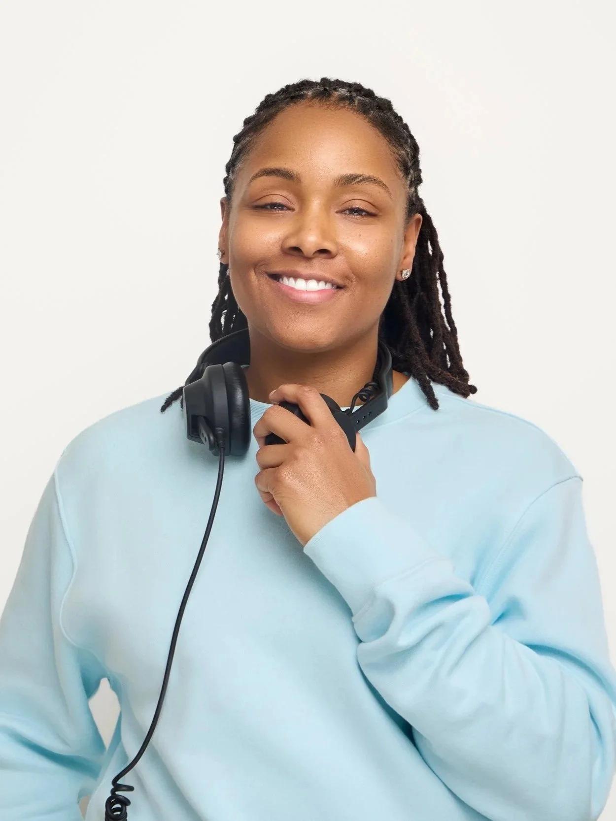 DJ Rhated R (Rhasheeda) of Chicago's Rhated Media Group smiling in front of a white wall, wearing headphones around her neck and a casual baby blue crewneck