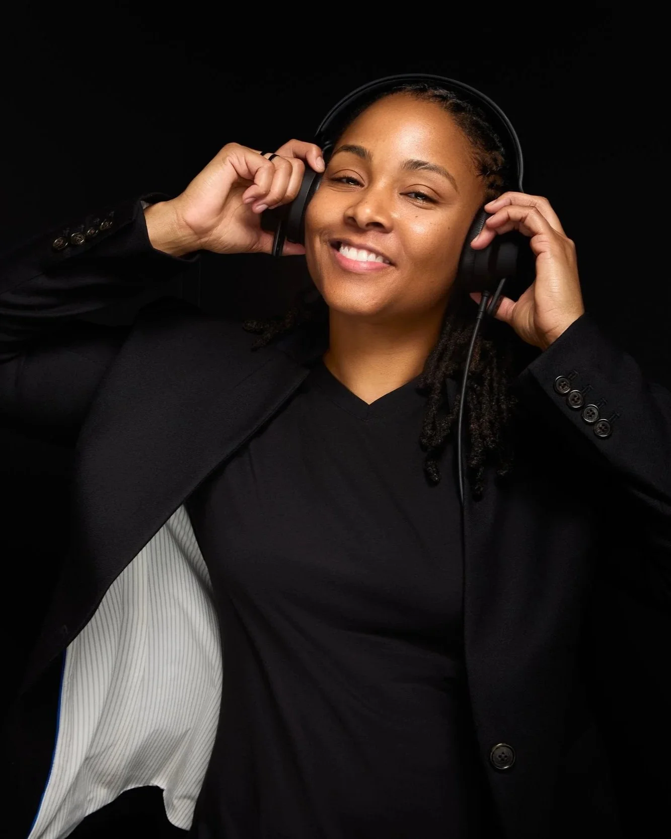 DJ Rhated R (Rhasheeda) of Rhated Media Group in Chicagoland, smiling in front of a black studio backdrop, wearing a black blazer and headphones while dancing