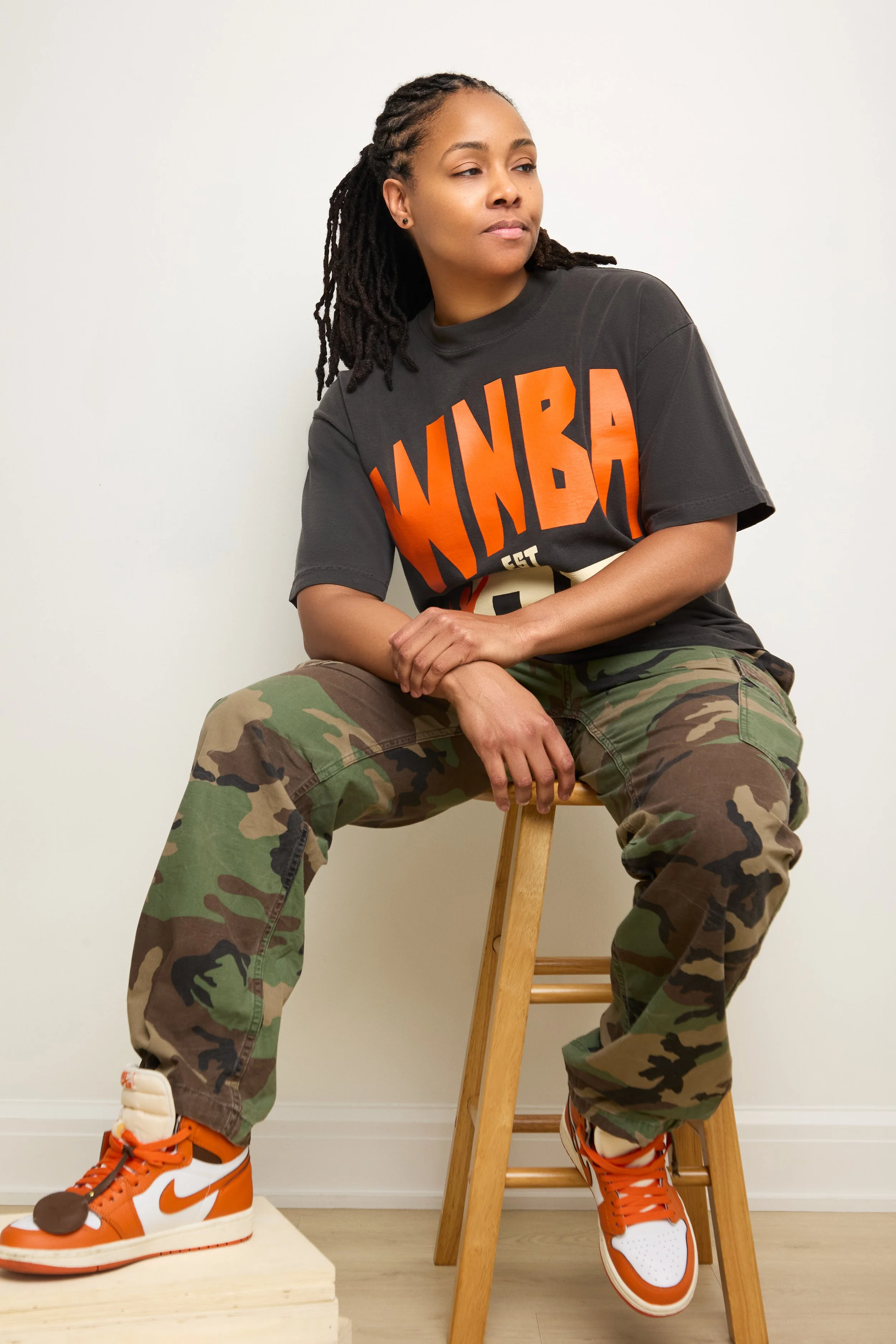 Chicago's DJ Rhated R (Rhasheeda) of Rhated Media Group sitting on a stool in front of a white wall, wearing a WNBA t-shirt, camo print pants, and orange Nike shoes