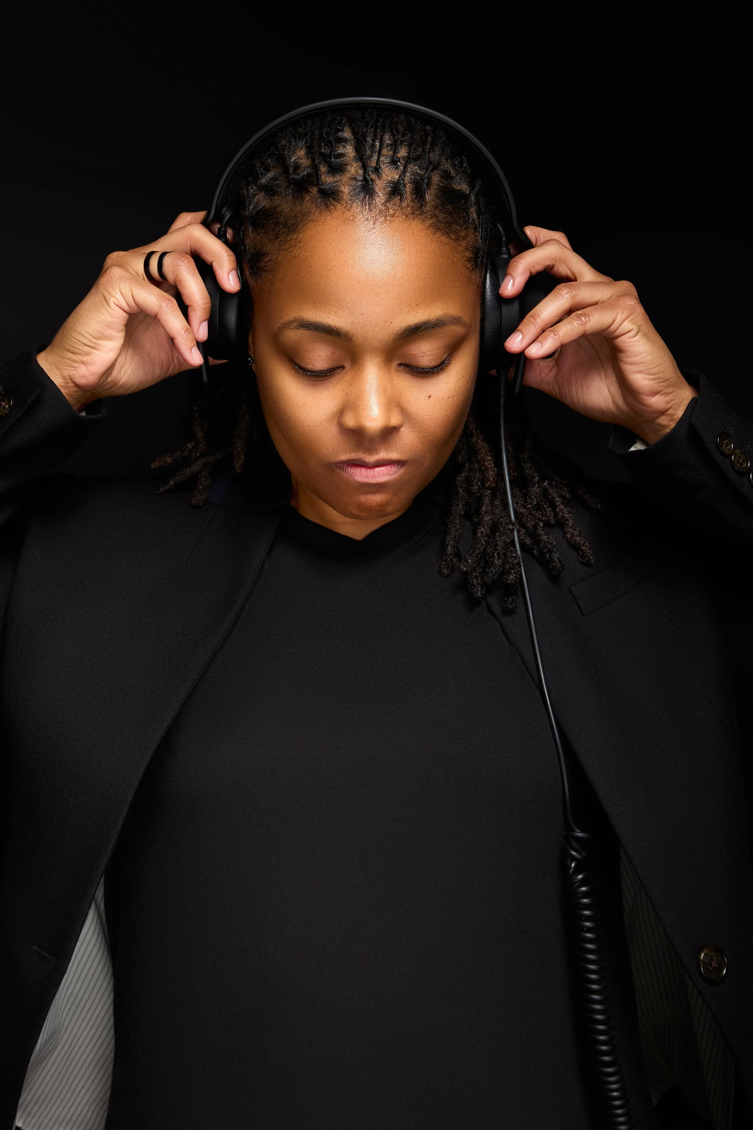 DJ Rhated R (Rhasheeda) of Rhated Media Group in Chicagoland, posing with a serious face in front of a black studio backdrop, wearing a black blazer and top, holding headphones while looking down towards a DJ table