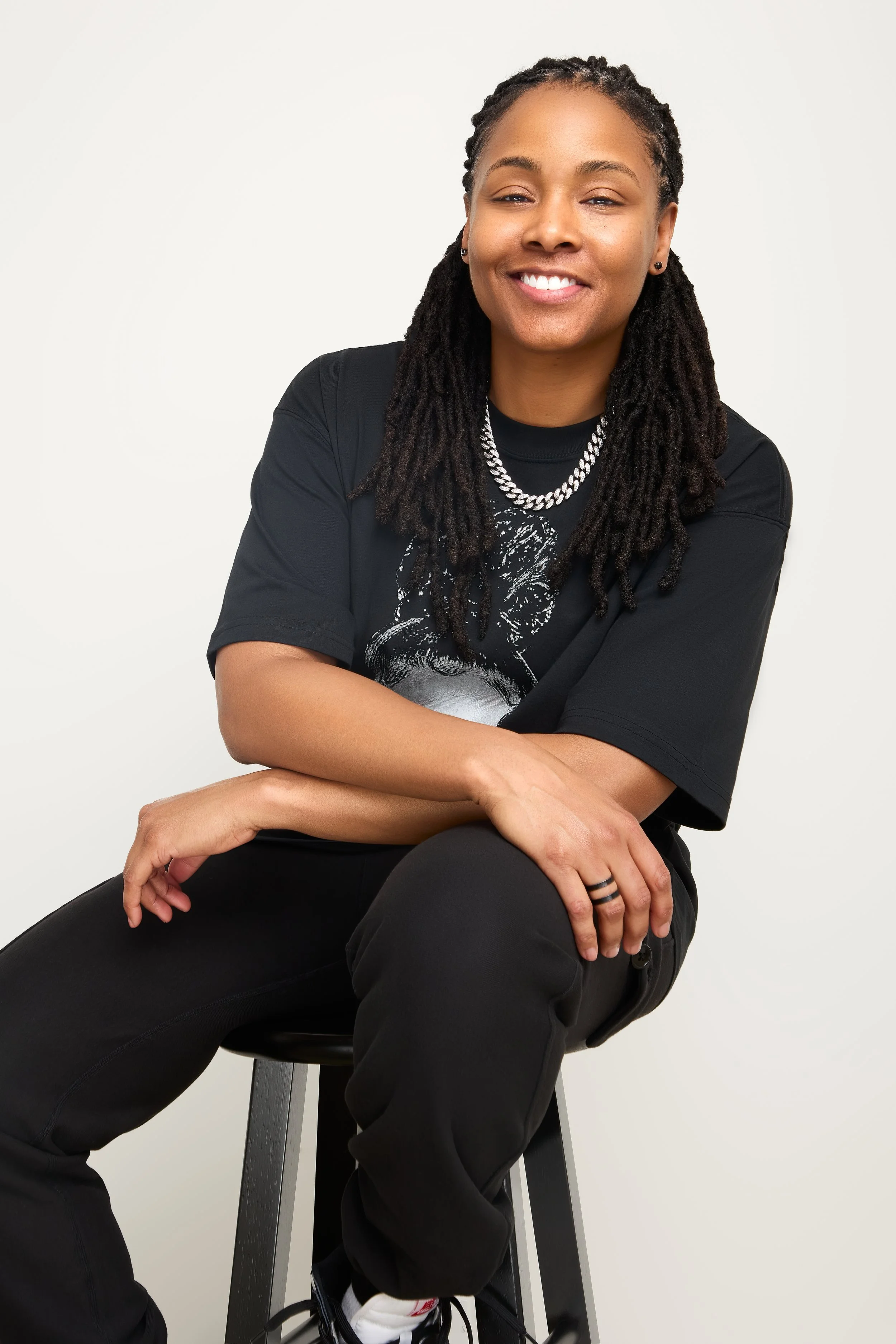 DJ Rhated R (Rhasheeda) of Rhated Media Group in Chicago, sitting on a stool in front of a white backdrop, wearing a black graphic t-shirt and pants, and a silver chain necklace, smiling at the camera