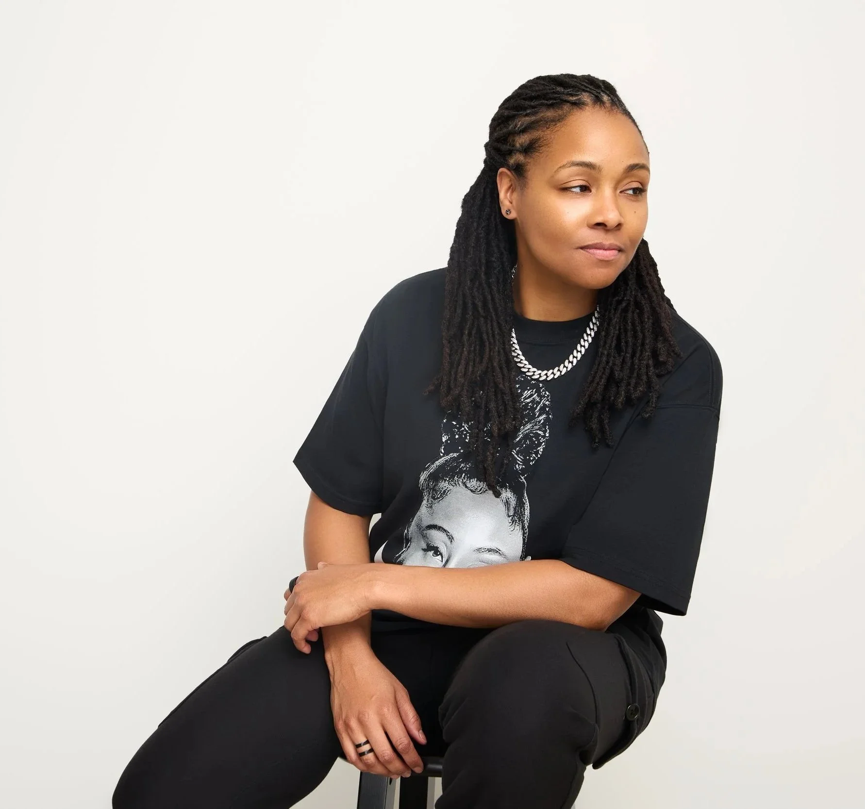 DJ Rhated R (Rhasheeda) of Chicagoland's Rhated Media Group, sitting on a stool in front of a white backdrop, wearing a black graphic t-shirt and pants, and a silver chain necklace, staring calmly to the right