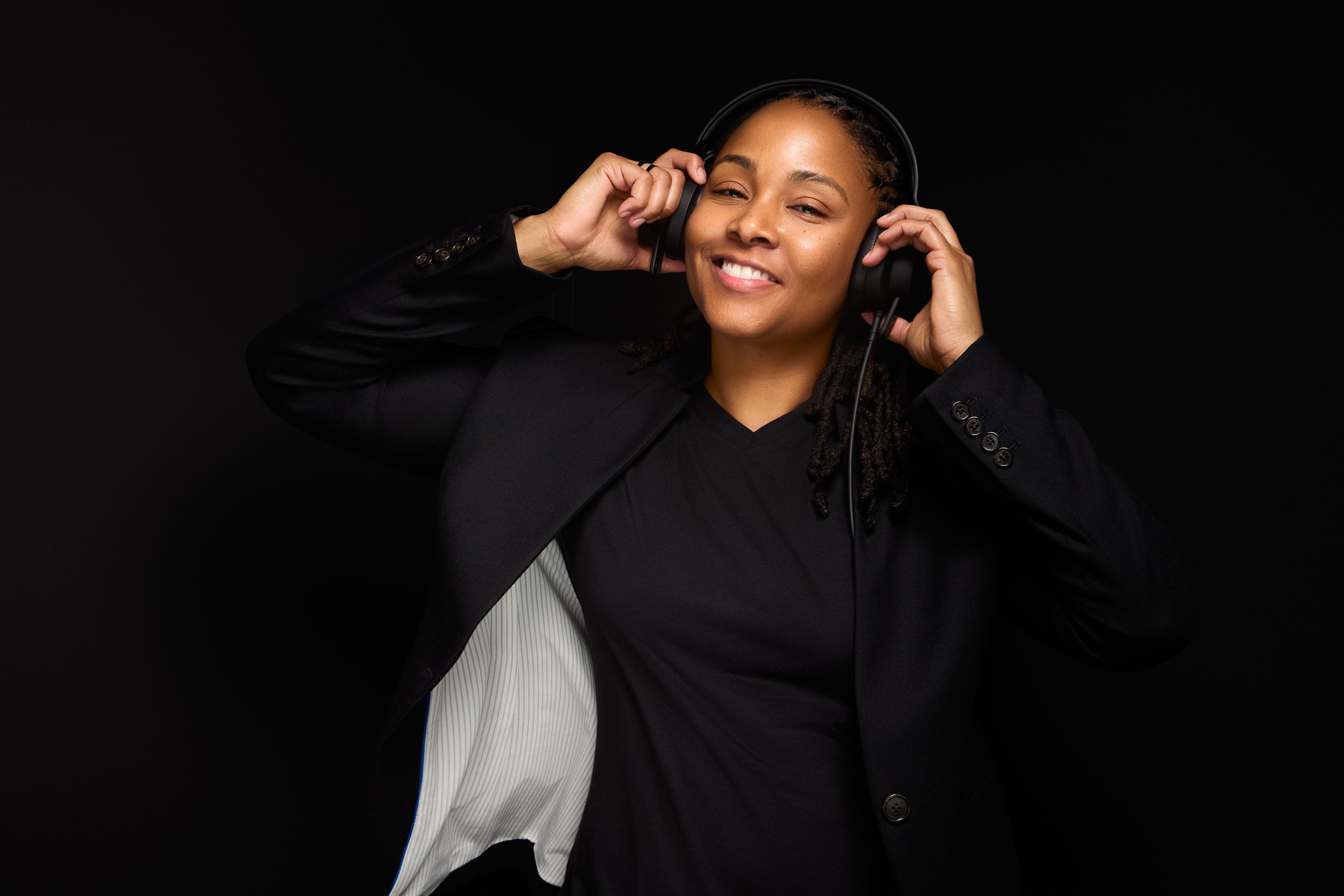 DJ Rhated R (Rhasheeda) smiling and dancing with headphones on in front of a black backdrop