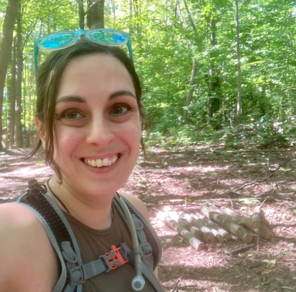 A white woman in a hydration vest on a trail in the wood smiling at the camera.
