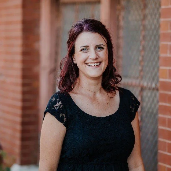 A white woman with her hair swept back into a half up, half down style, wearing a dark dress with an industrial brick background.