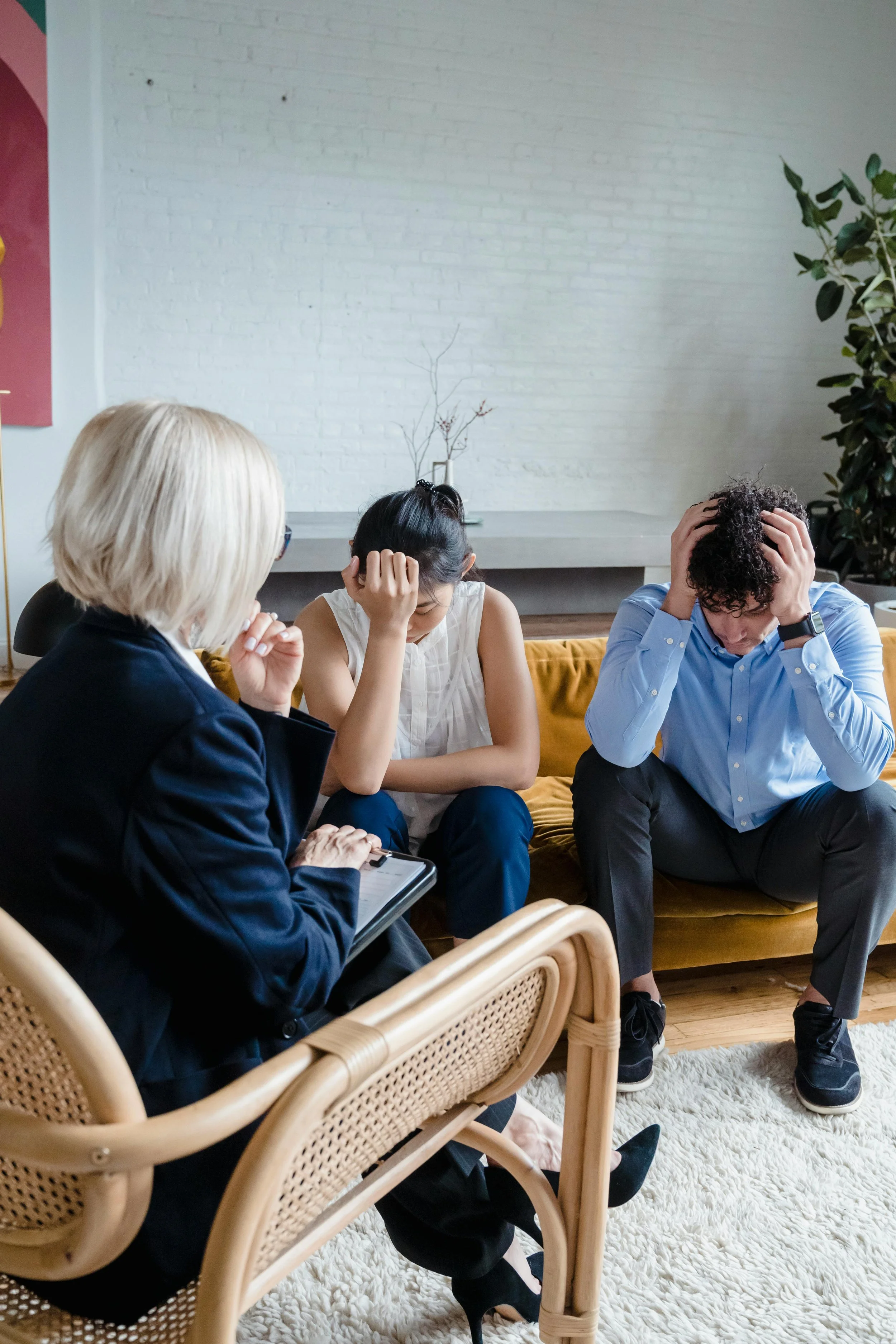 Three people sitting on a yellow sofa in a room with white brick wall, two women and one man, all appearing stressed or worried, with one woman taking notes on a notepad, the other woman holding her head, and the man holding his head with both hands.