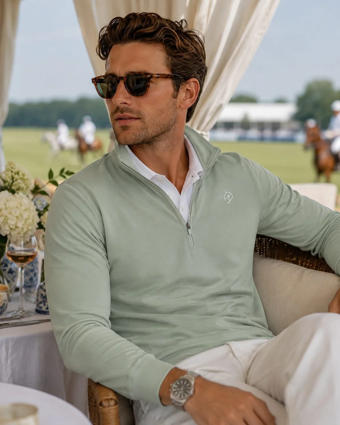 Man wearing sunglasses and a light green quarter-zip polo shirt sitting outdoors at a social event, with a table of flowers and drinks nearby, and horses and a tent in the background.