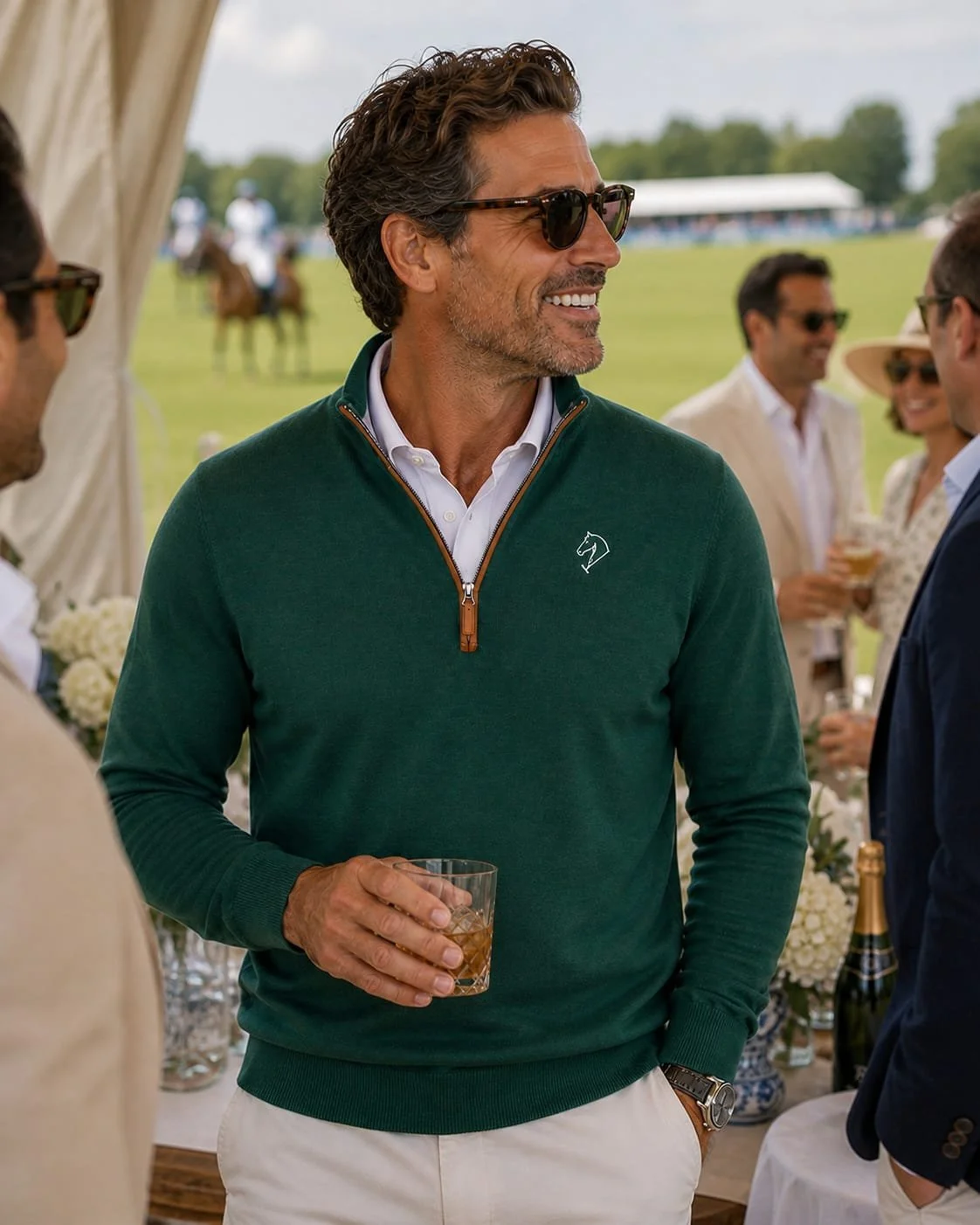 A man with sunglasses wearing a green quarter-zip sweater, holding a glass of whiskey, at an outdoor social gathering.