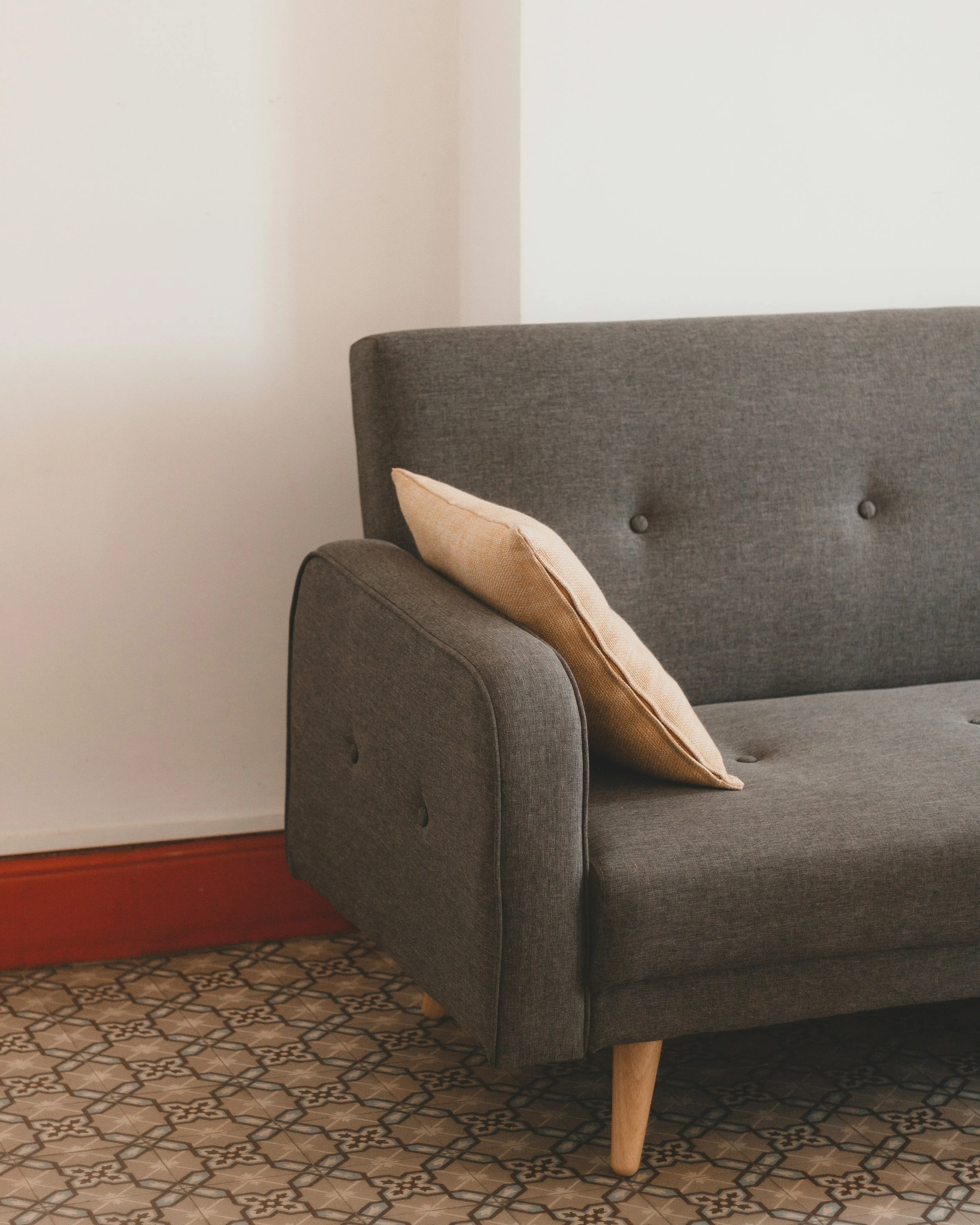Close-up of a dark gray fabric sofa with wood legs, beige throw pillow, set against a white wall with brown baseboard, on a patterned tile floor.