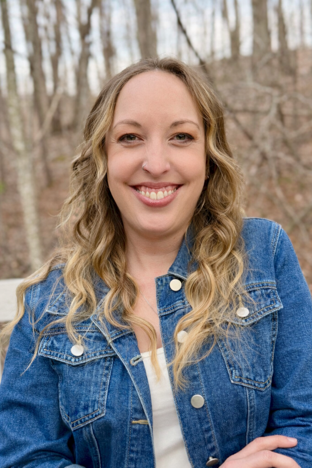 A woman with wavy blonde hair smiling outdoors in a wooded area, wearing a denim jacket.