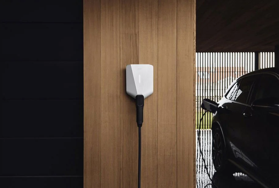 Electric vehicle charger mounted on a wooden wall next to a black car in a covered parking area.