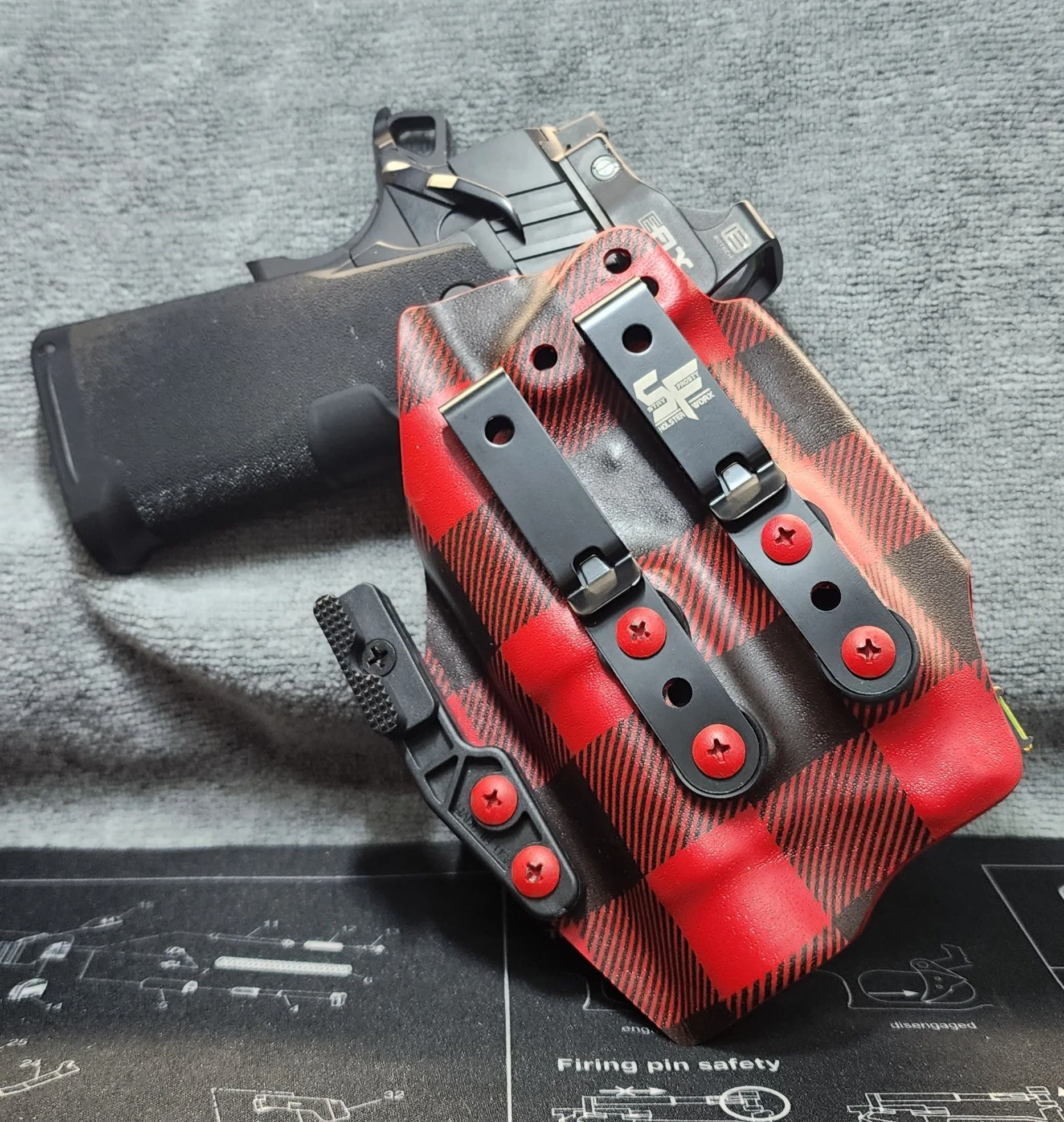 A handgun with a black grip and slide, resting on a gray surface, partly covered by a red and black plaid-patterned gun holster with black clips.