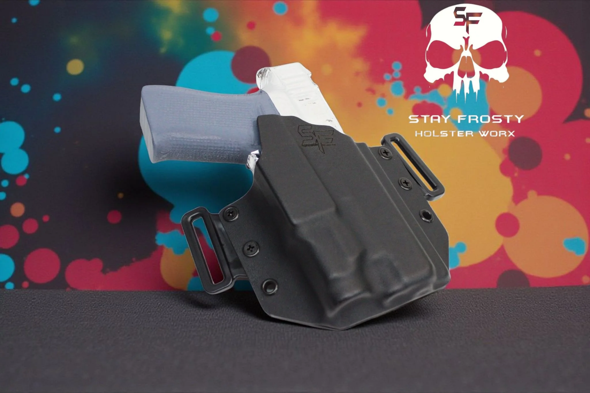 A black gun holster with a gray handle and a white attachment, placed on a colorful background with graffiti-style splashes of pink, orange, blue, and black. Text on the background reads 'Stay Frosty' and 'Holster Worx' with a white skull logo at the