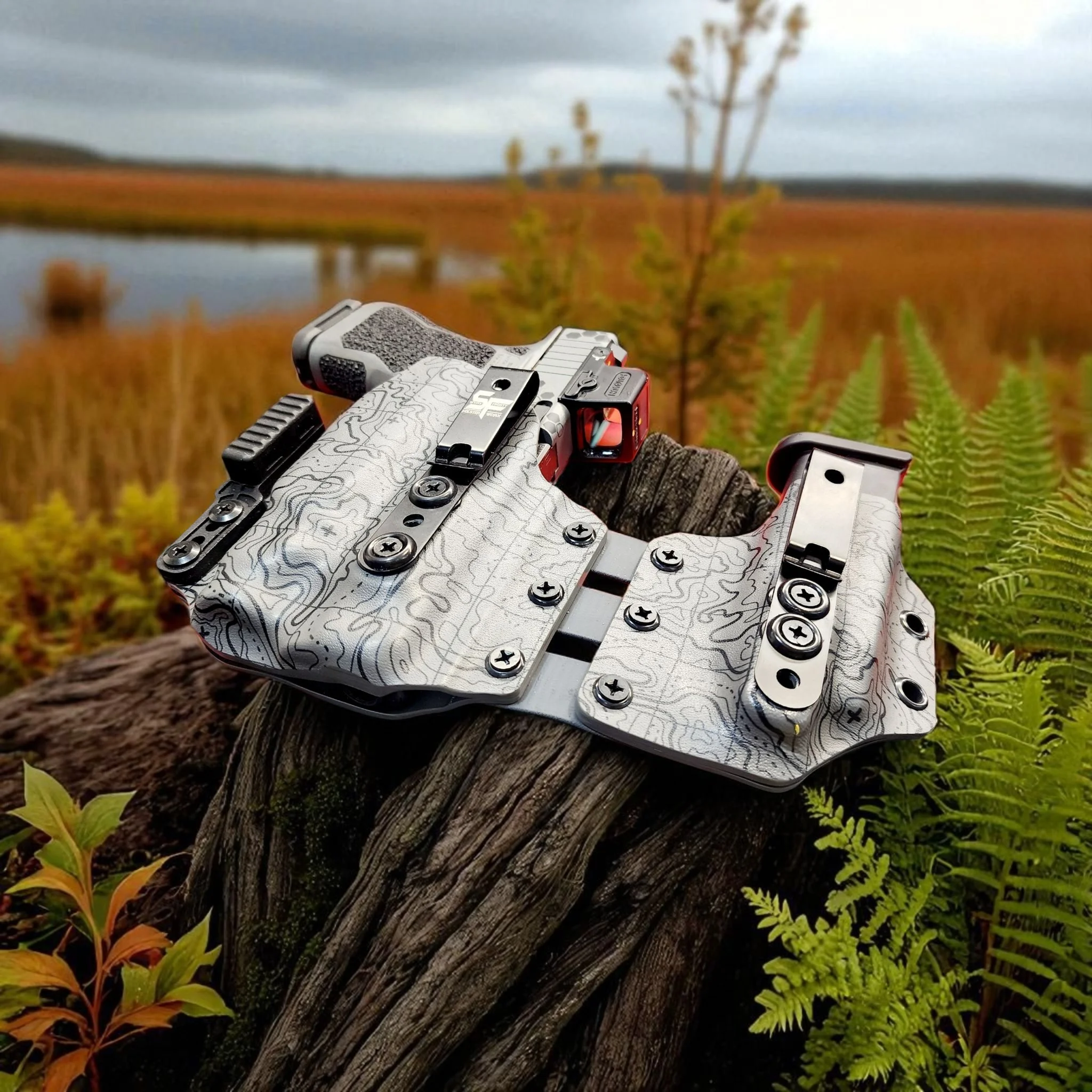 A gun with a topographical map design resting on a tree trunk outdoors in a landscape with ferns, grass, a small lake, and a cloudy sky.