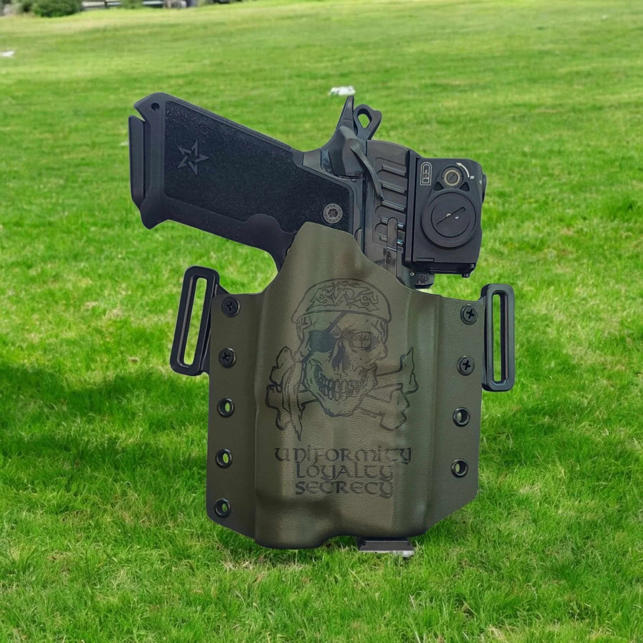 A handgun in a green tactical holster with a skull and lettering design, held upright in the grass.