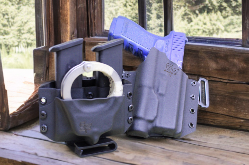 A handgun, a tactical flashlight, and a multi-tool knife in black Kydex holsters on a wooden surface near a window.