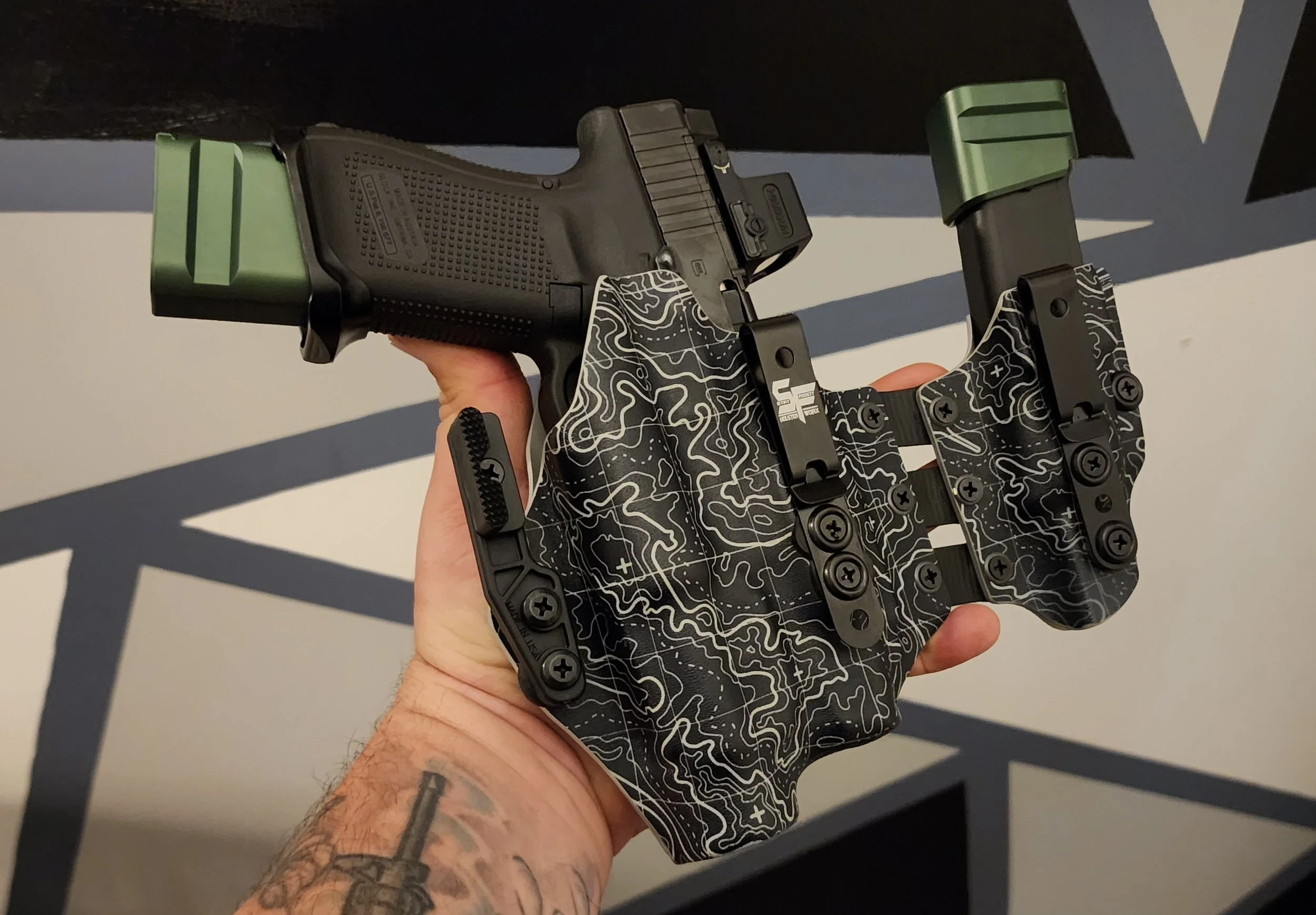 A handgun with a black grip and green slide, held by a person with a tattooed arm. The gun is mounted on a black and white patterned device with screws.