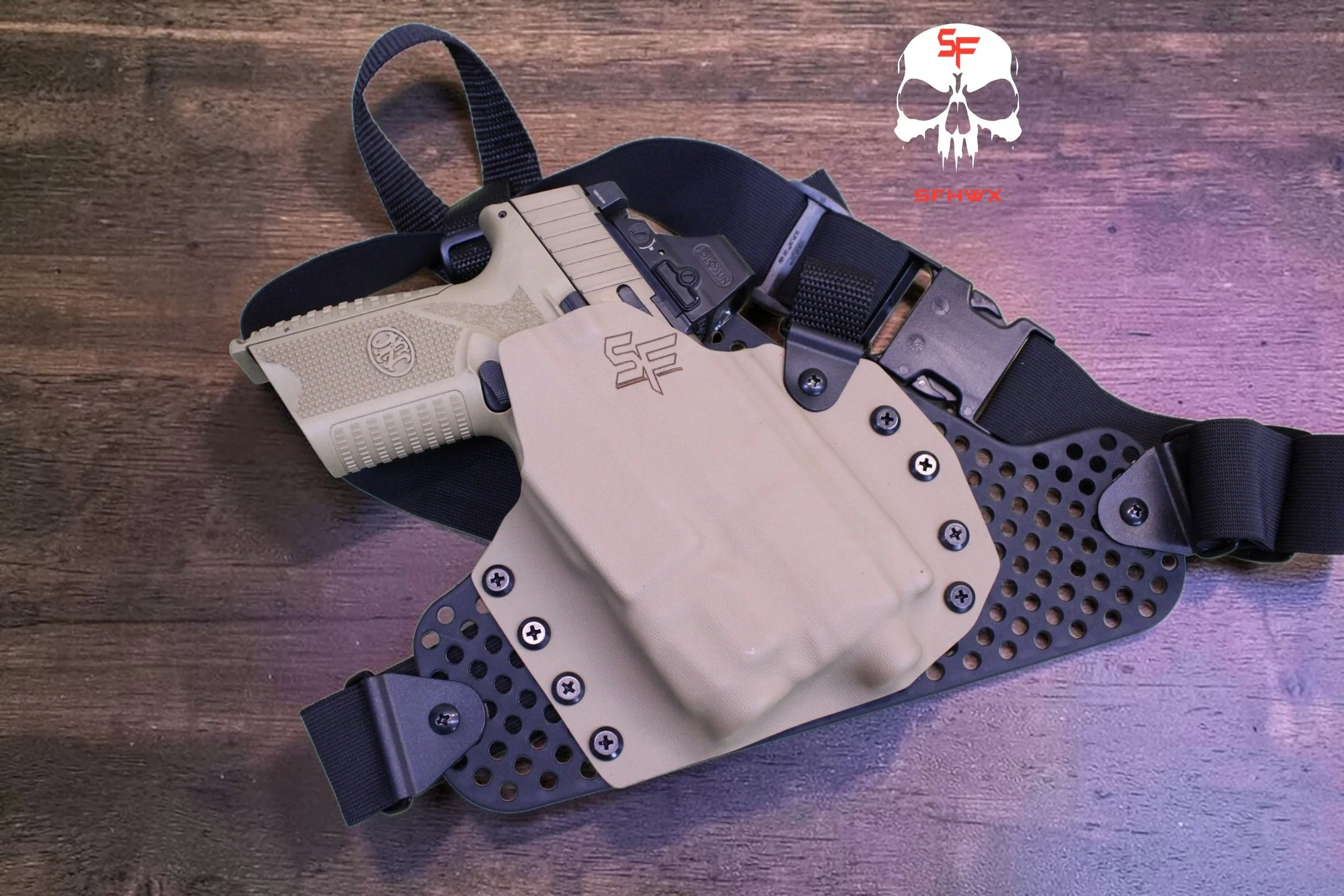 Tactical holster with a beige handgun inside, attached to a black harness on a wooden surface. The holster has several screws and a section with holes for ventilation, and there is a logo with the letters SF on the holster and a skull with red and wh