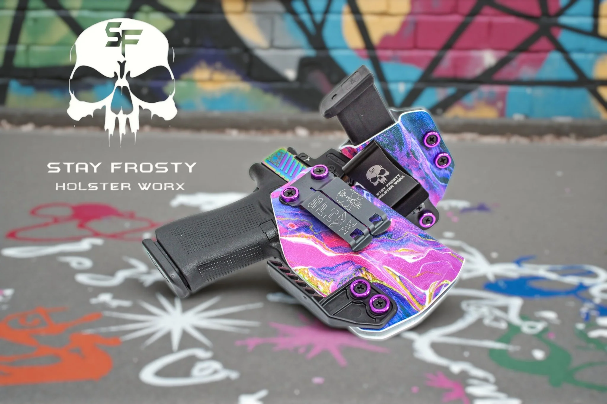 A handgun with a colorful marbled slide and purple screws, attached to a purple and blue gripped holster with skull motifs, placed on a concrete surface decorated with multicolored spray paint graffiti, with a graffiti-covered wall in the background.