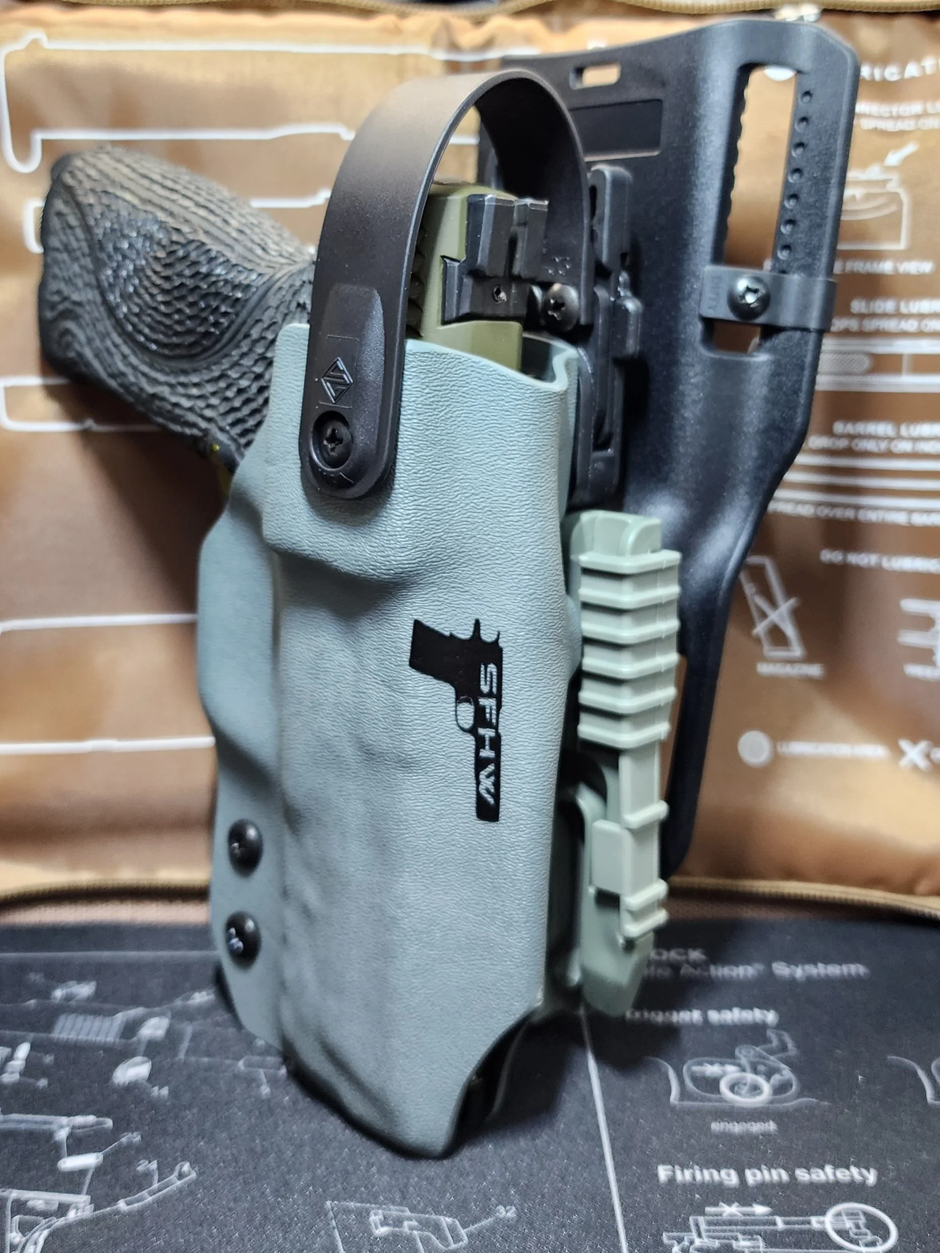 Black handgun trigger guard mounted on a robotic or tactical handgun with a light gray grip panel marked with a black gun logo and text.