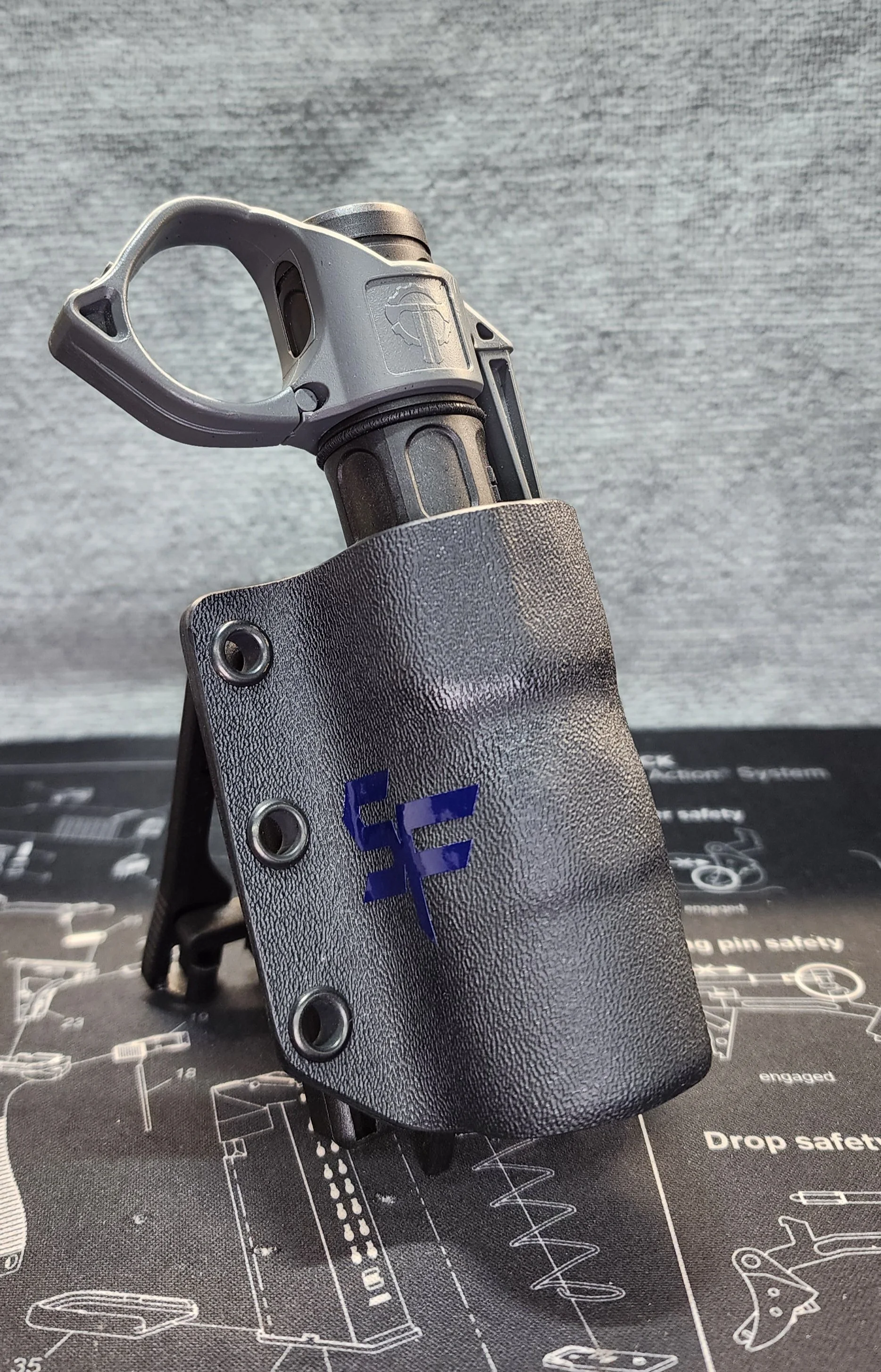 A close-up of a handgun in a black holster with a purple logo, placed on a black mat with technical diagrams and text.