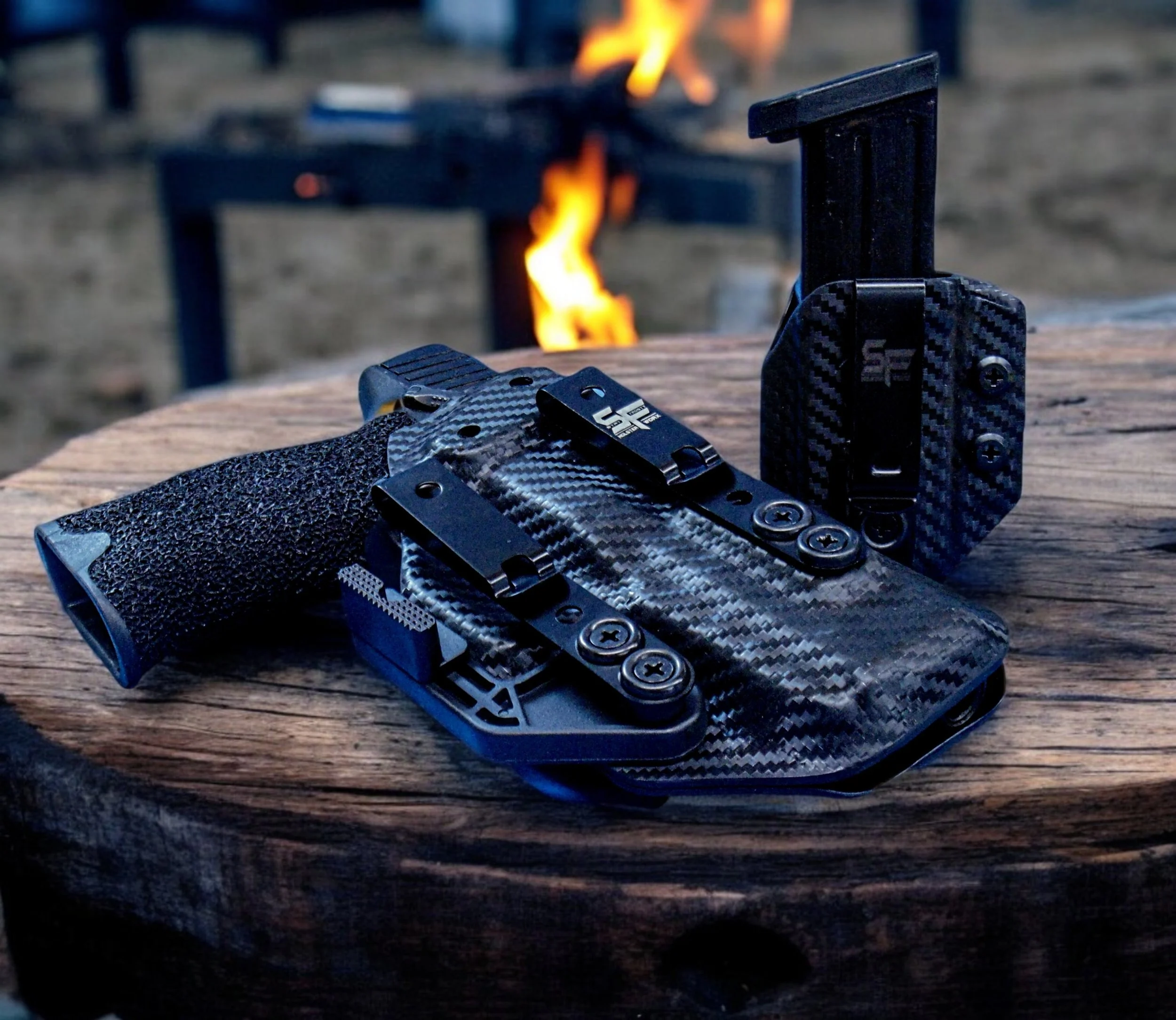 Close-up of a black and gray tactical knife with a textured grip, placed on a wooden surface with a blurred outdoor background featuring a campfire.