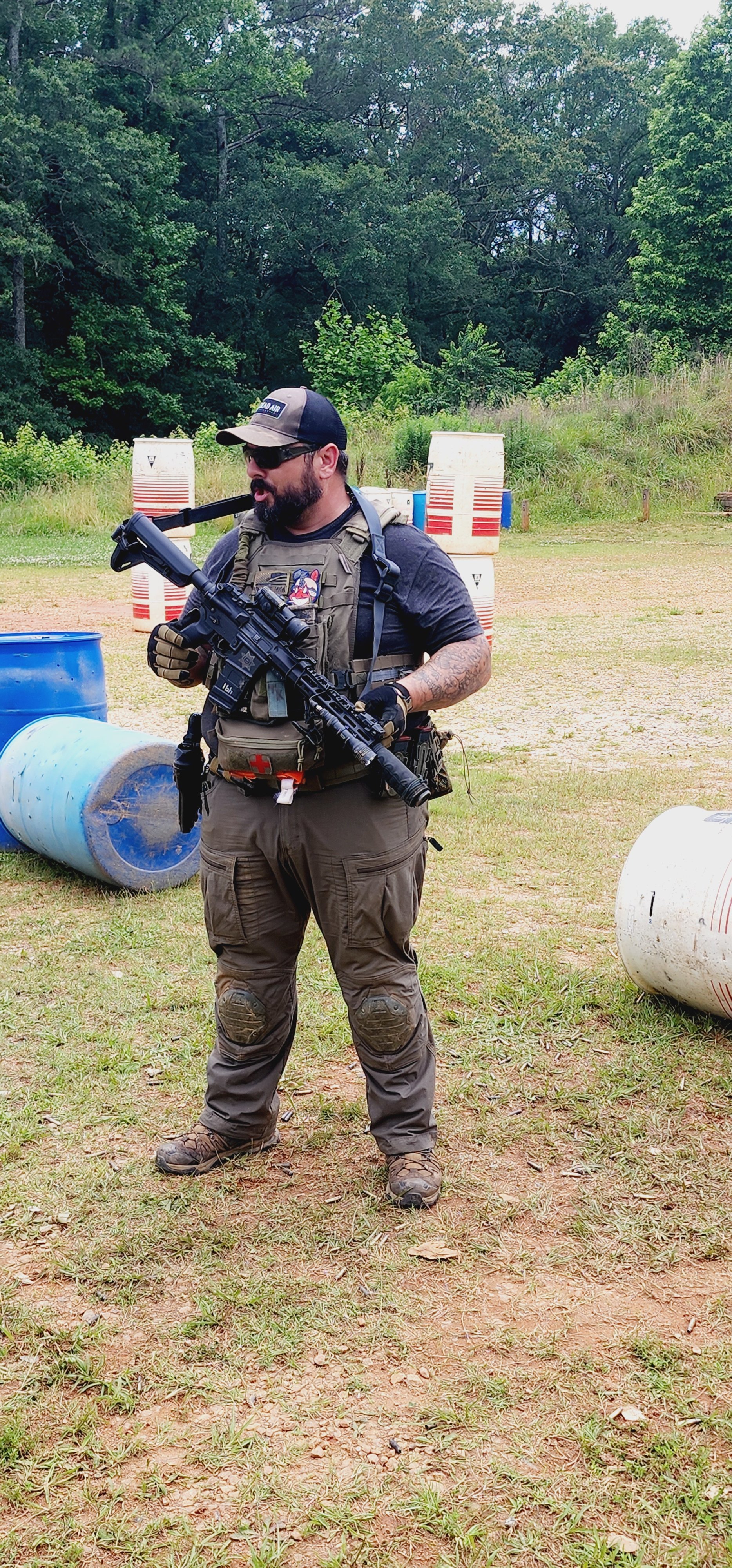 Man dressed in tactical gear standing outdoors on a grassy field, holding a rifle, with trees and large barrels in the background.