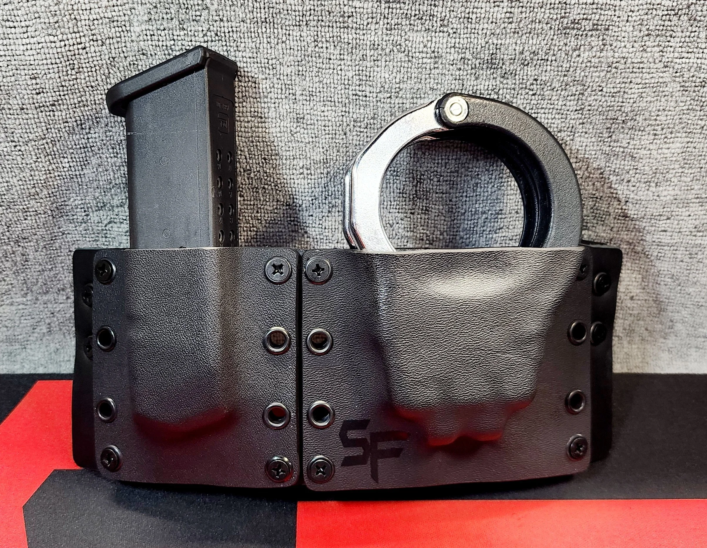 A black tactical belt with a large, round metal buckle, attached to a black holster with screws and a magazine holder on a gray textured surface.