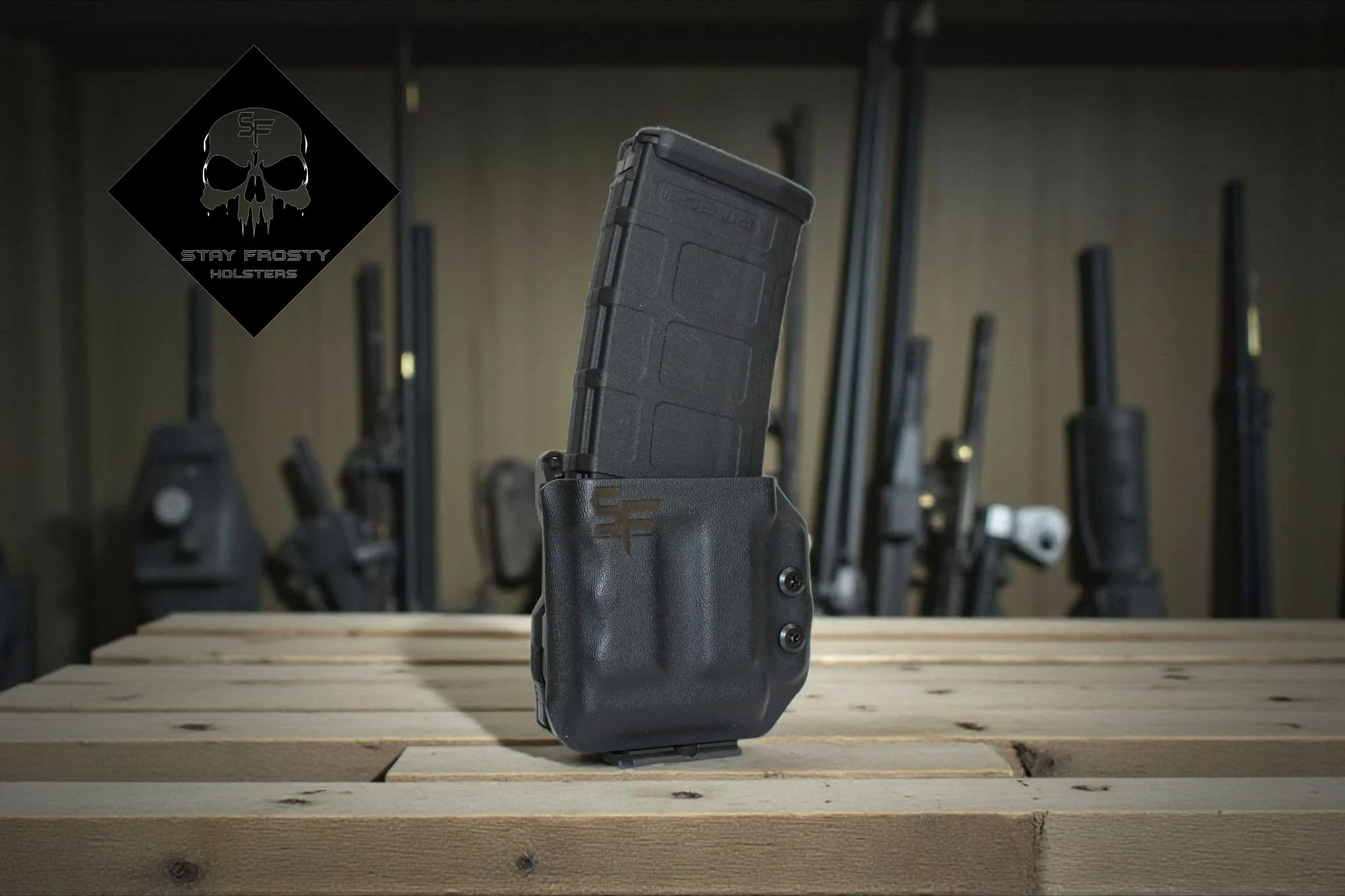 A handgun magazine loaded with ammunition inside a black holster, placed on a wooden surface. In the background, there are multiple gun holsters mounted on the wall. A logo with a skull and the text 'Stay Frosty Holsters' is visible in the upper left