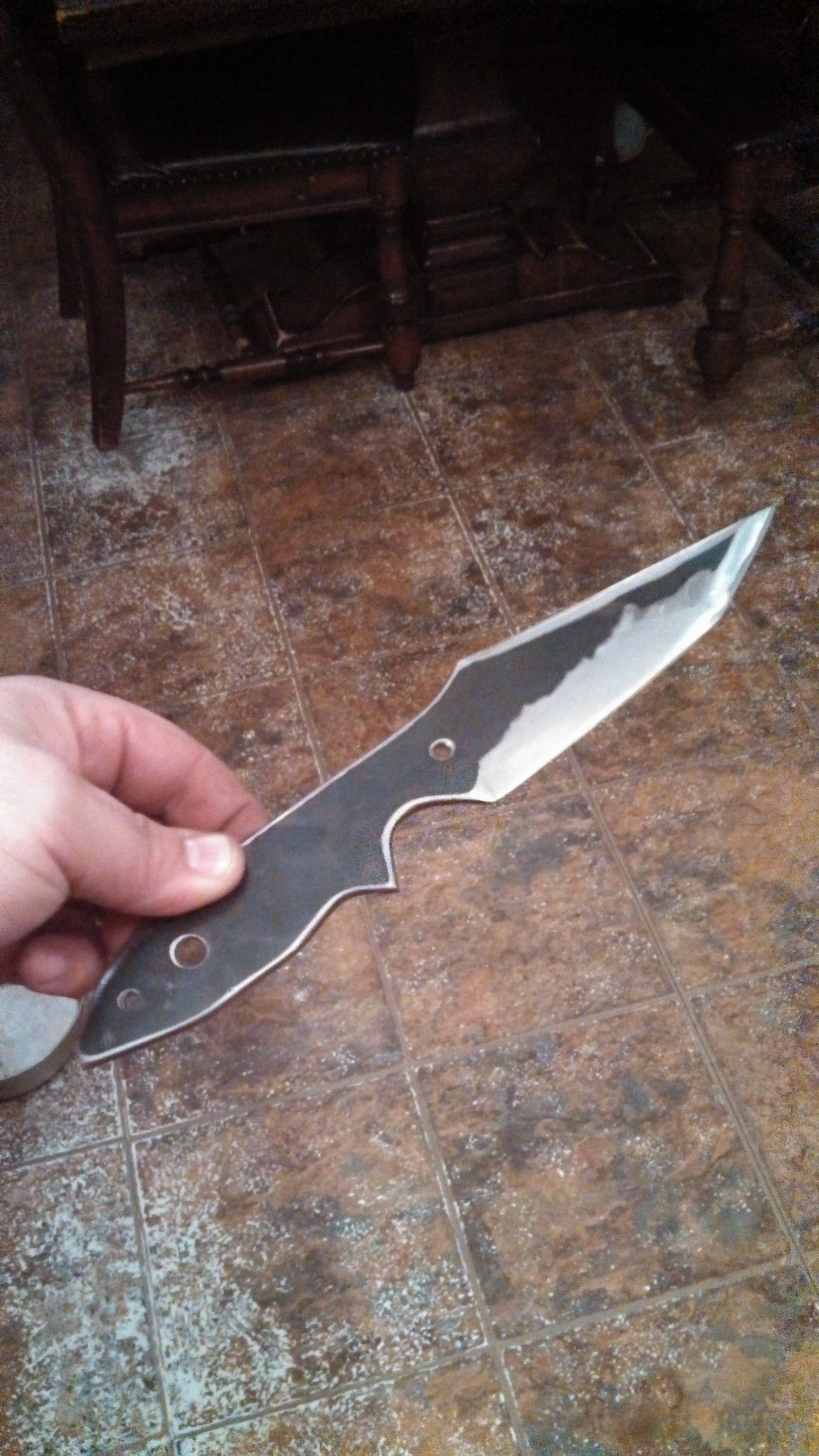 Person holding a partially finished knife with a plain handle, pointed blade, and holes in the handle for attachment, against a tiled floor and wooden furniture in the background.